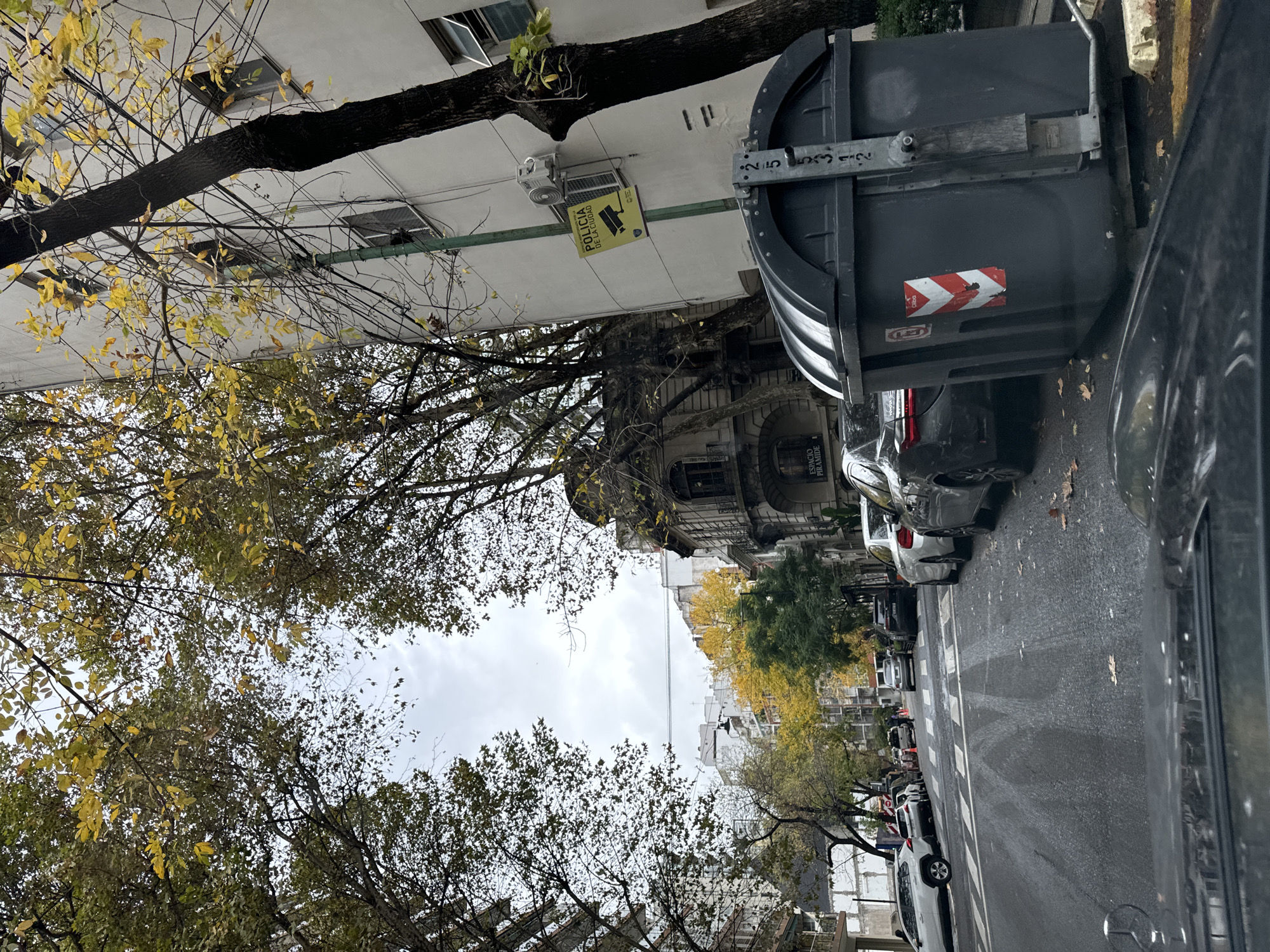 The image captures a street scene in Buenos Aires, Argentina, characterized by autumn foliage. Trees with yellowing leaves line the street, framing the view towards the sky. Buildings with architectural details are visible along the street. A dumpster is positioned on the sidewalk and several cars are parked along the curb. A sign indicating "Policia de la Ciudad" (City Police) with a surveillance camera icon is mounted on a building. The sky is overcast.