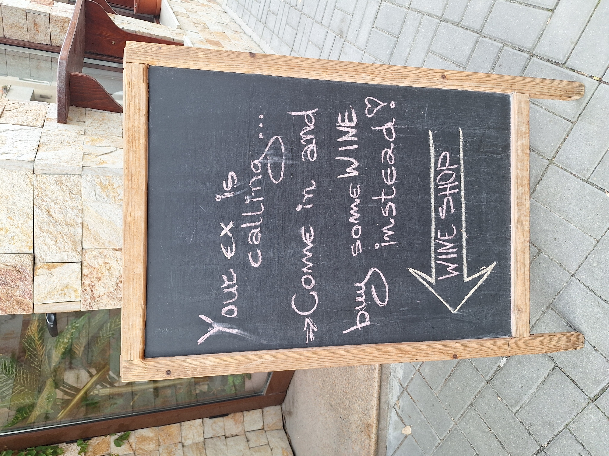 This eye-level outdoor shot depicts a chalkboard sign propped against a building on a paved area. The sign, framed in light-colored wood, features a handwritten message in pink chalk: "Your ex is calling... ⇒ Come in and buy some WINE instead!," with an arrow pointing down to the words "WINE SHOP."
The sign's right corner is situated on a gray-paved surface, likely a sidewalk. The sign's left corner is positioned against a textured beige stone building with an adjacent window containing foliage. Above the window, a small brown wooden shelf is visible. The location is in the city of Varna, Bulgaria.