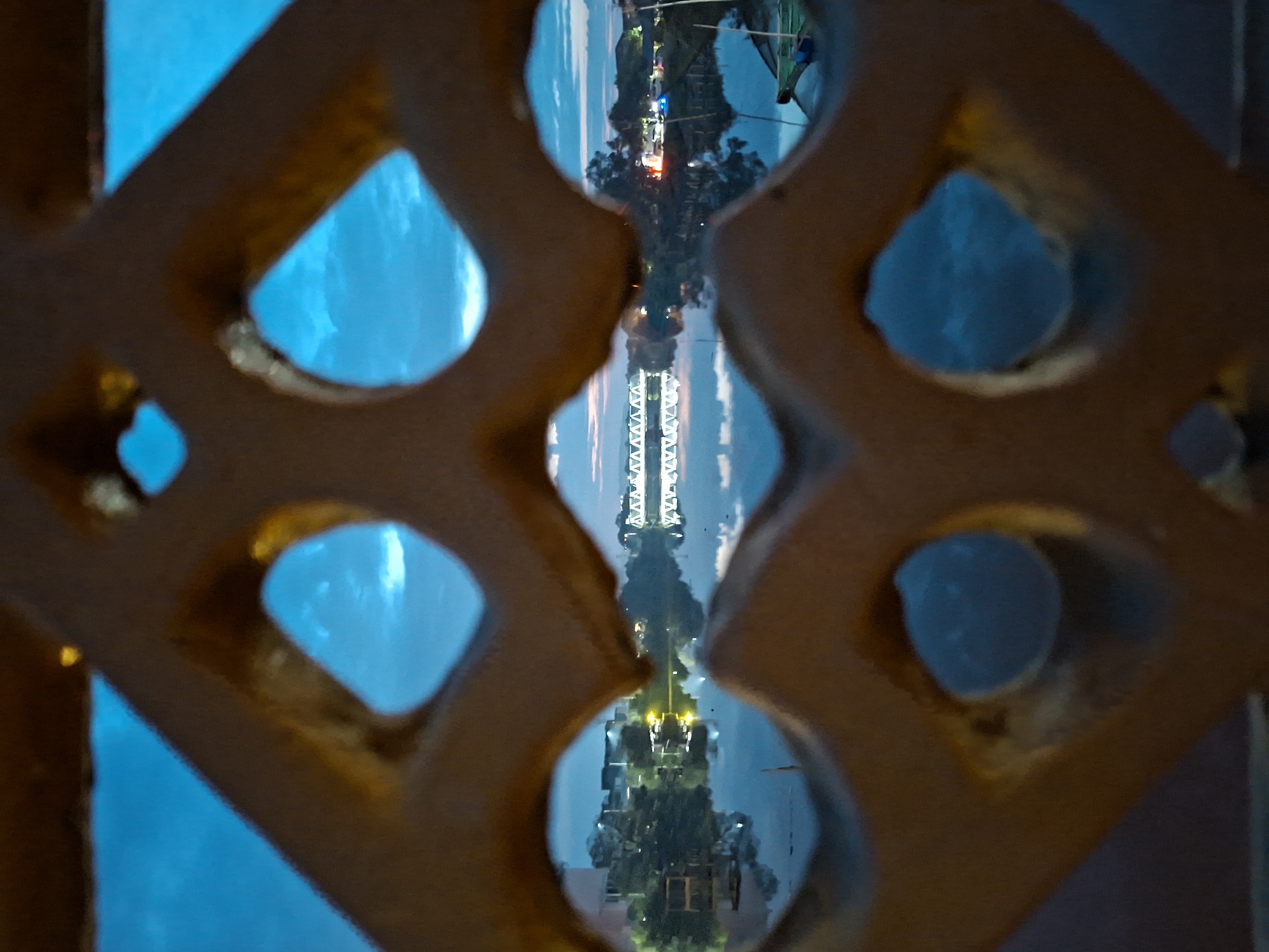 The image is a photograph taken from behind a decorative, possibly metal, screen with a geometric, lattice-like pattern. The screen is in the foreground and is out of focus, appearing in shades of brown and gold. Through the openings of this screen, a serene twilight scene of a body of water, likely a river or bay, is visible. The sky is a deep, twilight blue with hints of lighter clouds.
On the far side of the water, a bridge with a distinctive truss structure is illuminated, its reflection shimmering on the calm water's surface. To the left and right of the bridge, the distant shore is lined with trees and buildings, some of which are lit by artificial lights, suggesting a town or city. The overall atmosphere is tranquil and atmospheric, with the interplay of light and shadow characteristic of dusk. The scene appears to be located in Lhokseumawe, Indonesia, as indicated by the context. There are no people clearly visible in the image. The water is still, reflecting the sky and the lights on the shore. A small boat with a green hull and a white pole is partially visible on the right side of the image, floating on the water.