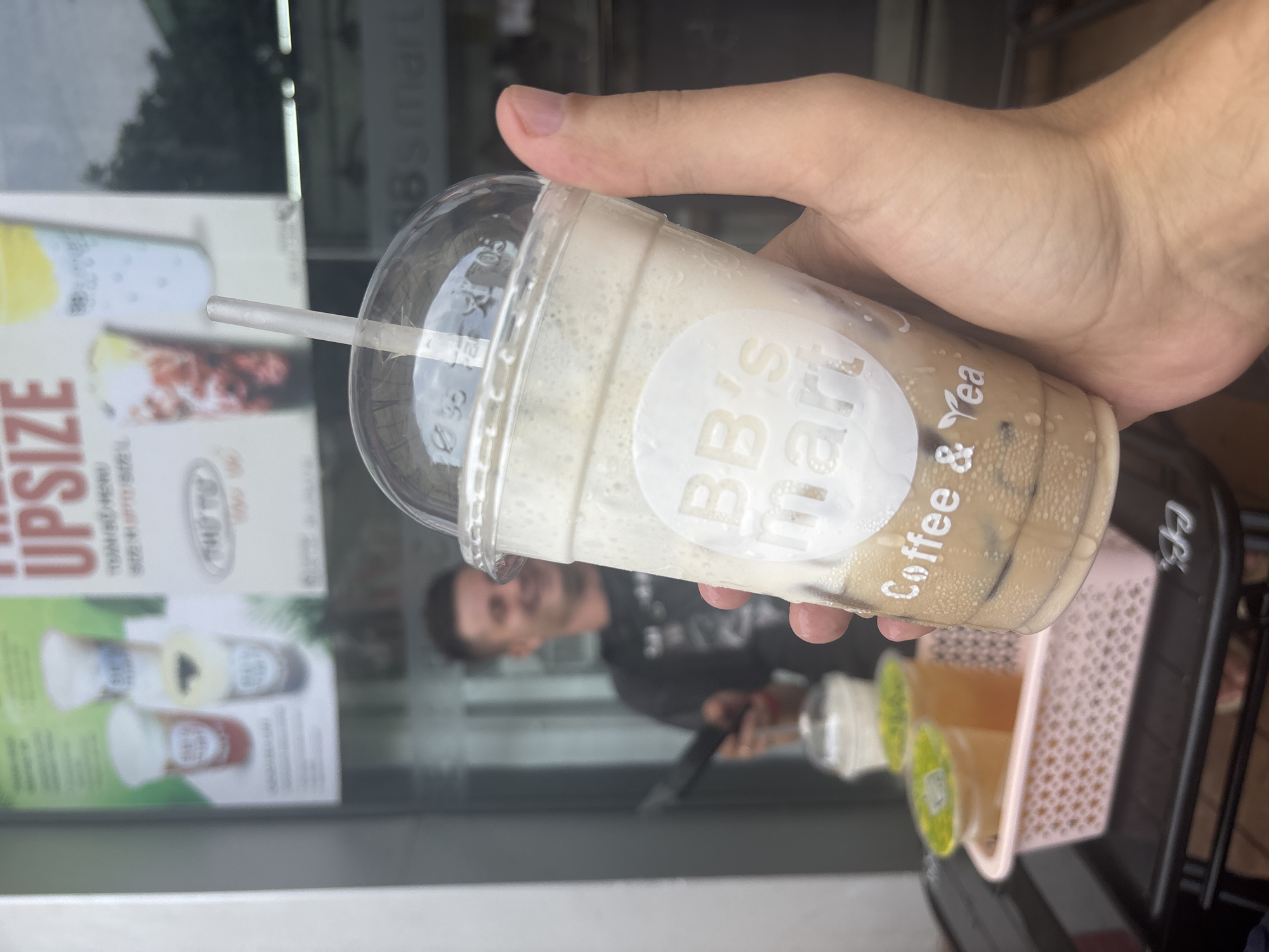 An outdoor, eye-level, medium shot captures a person's hand holding a clear plastic cup filled with a layered coffee beverage, likely an iced latte or frappe, with a white, foamy top and darker liquid with ice at the bottom. The cup is adorned with a circular white sticker that reads "BB's mart Coffee & Tea." A thin, clear straw protrudes from the cup's plastic lid. In the blurred background, a smiling man in a black t-shirt is visible, seemingly working behind a counter or window. To the left, a pink plastic basket holds three other clear plastic cups with drinks, one with a green lid and two with yellow lids. The setting appears to be a cafe or street vendor stall.