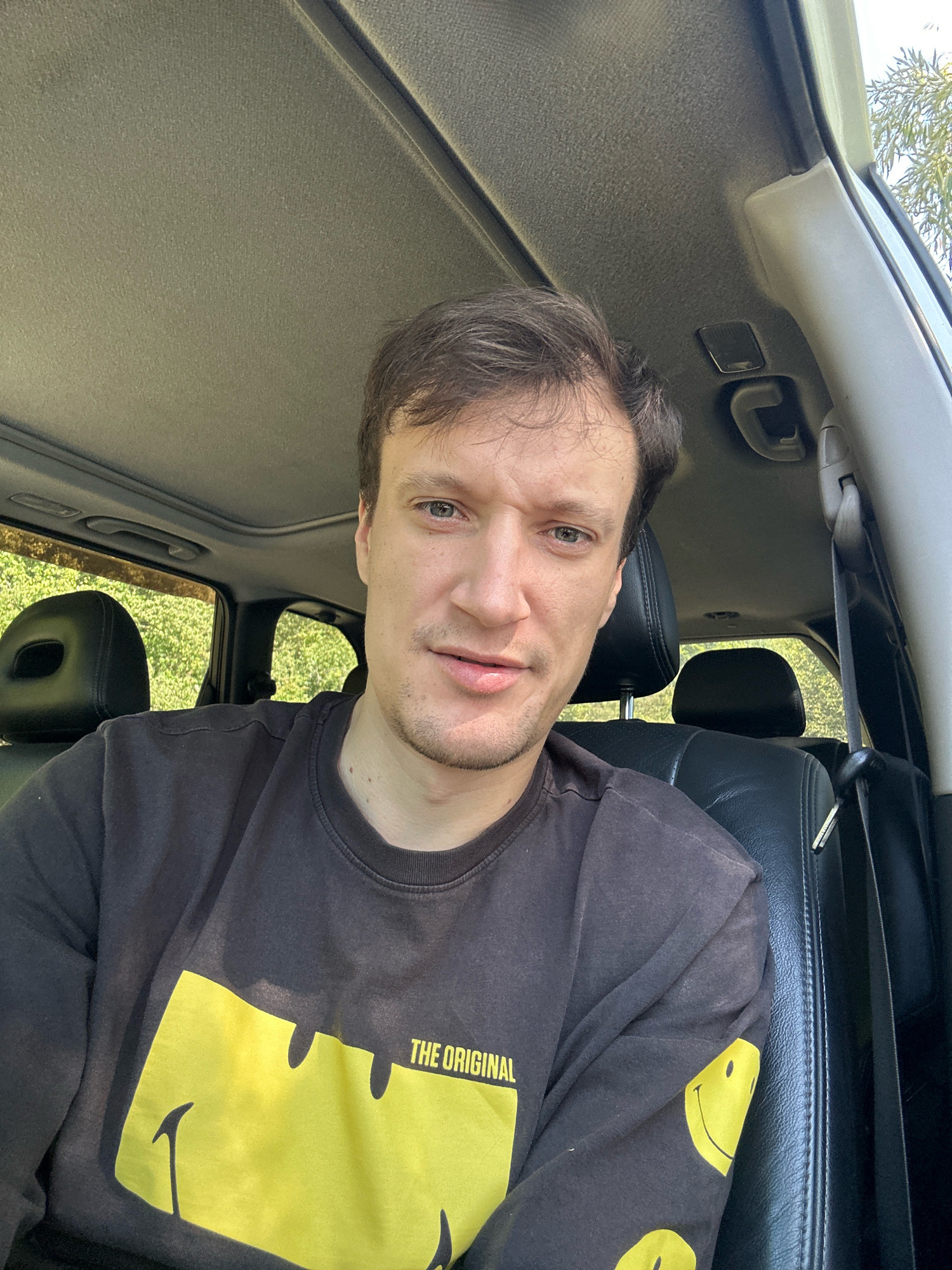 A young man is sitting in the driver's seat of a car. He is wearing a dark long-sleeved shirt with a yellow smiley face graphic on it. The car's interior is visible, including black leather seats, the ceiling with a sunroof, and the front windshield. Outside the car, lush green foliage is visible, suggesting a rural or suburban setting. The man is looking directly at the camera, with a neutral expression. The scene appears to be a selfie taken inside a vehicle, possibly in a rural area near Makiv, Ukraine, given the natural, undeveloped surroundings.