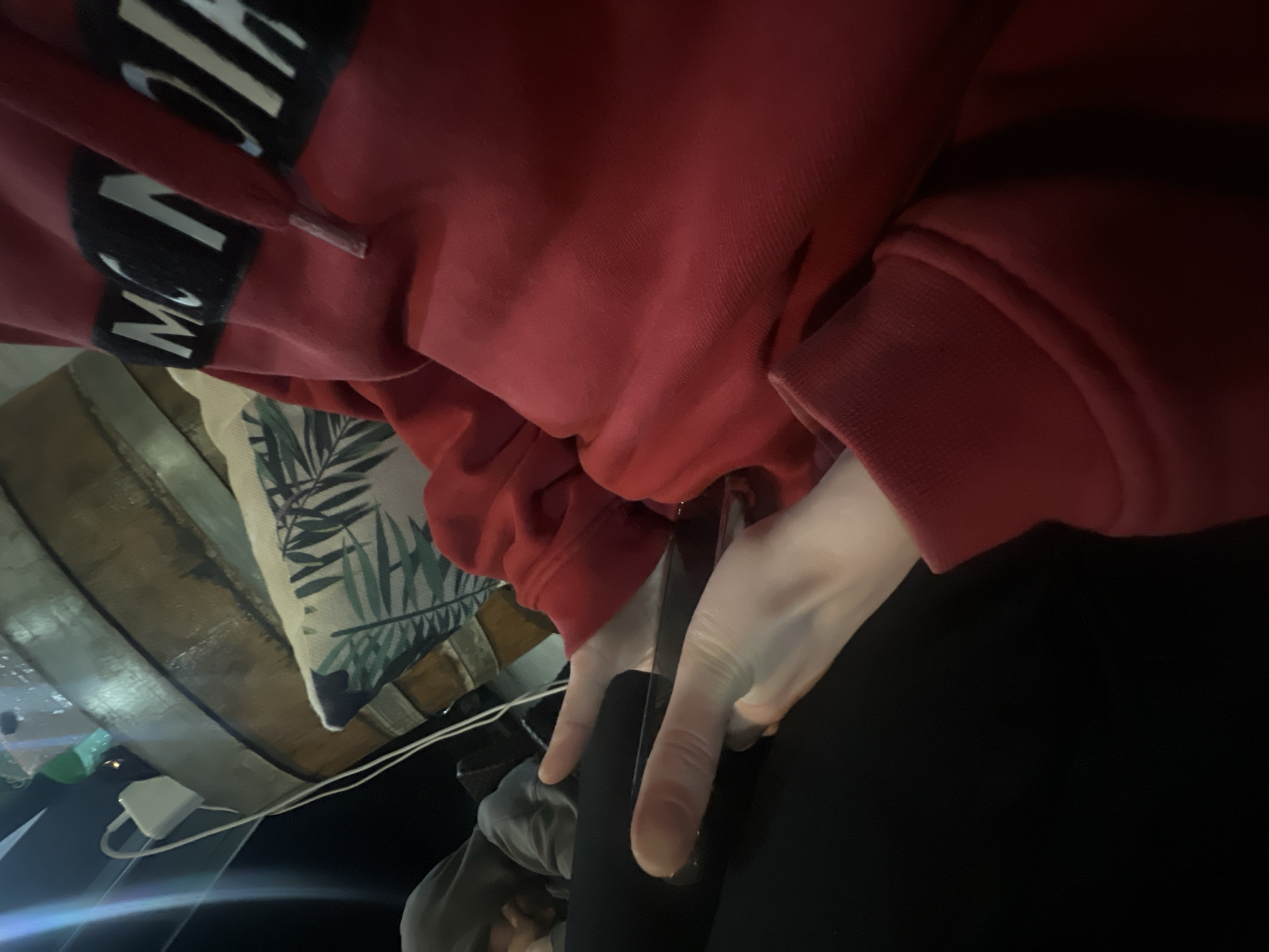 The image is a low-angle, indoor shot of a person wearing a red sweatshirt with the word "MONCLER" written on it in large, white capital letters against a black background on the front. The person is holding a black smartphone in their hands and wearing black leggings. The background includes a wooden surface, a pillow with a green and white leaf pattern, a white charging block, and a cable. The location is unknown.