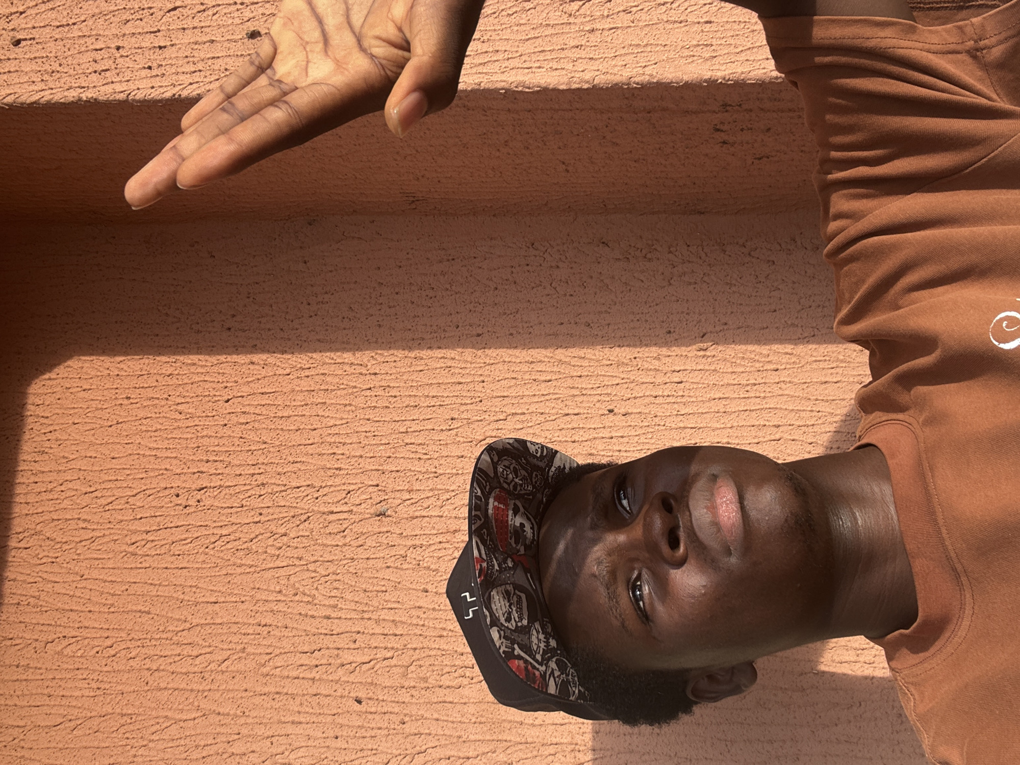 A young man is positioned in the lower right corner of the image, appearing to be outdoors with a textured, peach-colored wall as the background. The man is wearing a brown t-shirt and a black baseball cap adorned with skull and crossbone designs. His face is tilted upwards towards the camera, with his eyes open. Above the wall, at the upper edge of the image, is a hand with fingers extended. The setting is outdoors. The city of Onitsha, Nigeria cannot be explicitly verified through this photograph.