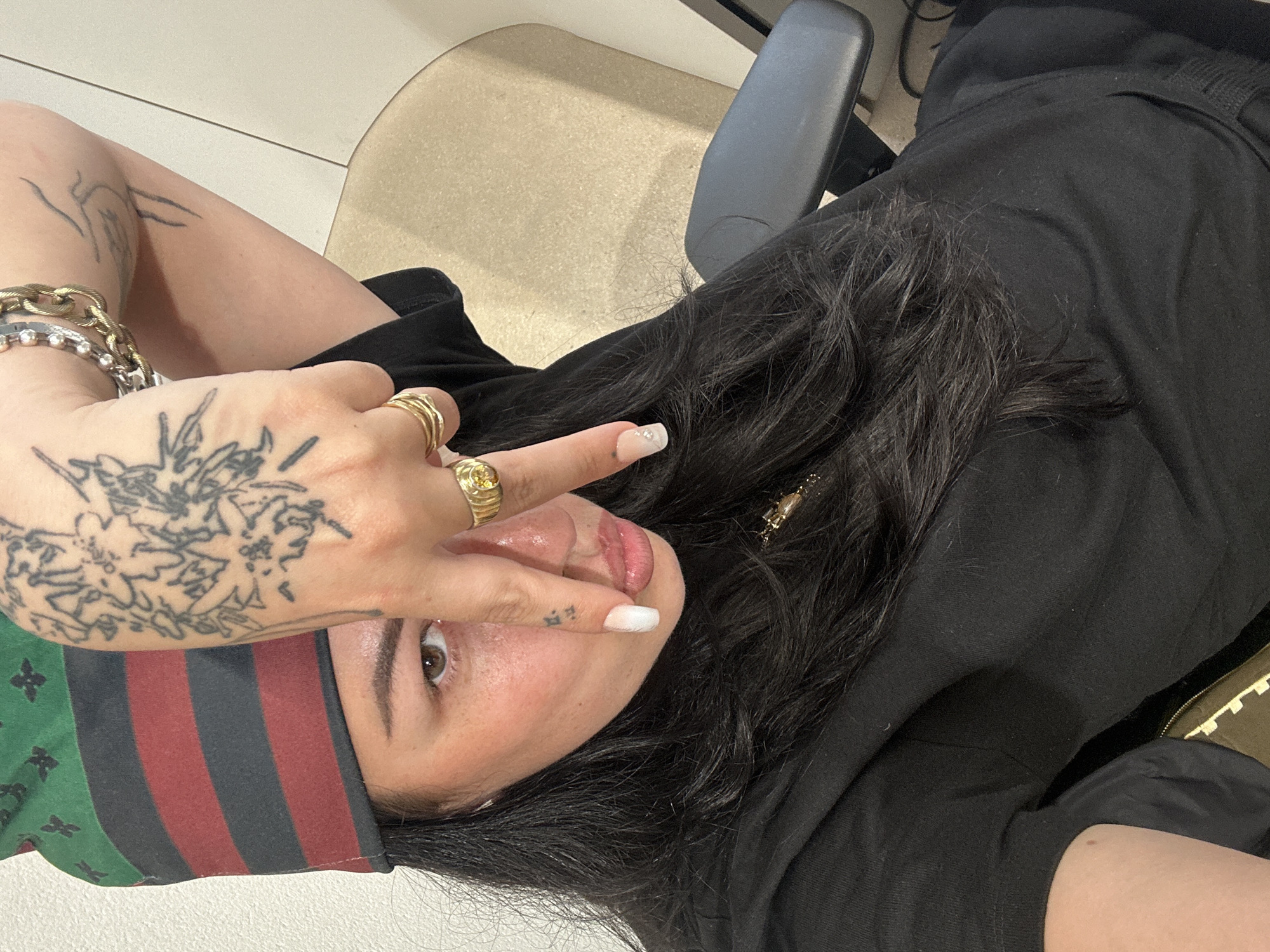 A person with multiple tattoos and wearing several rings is positioned in front of an office chair with a tan seat and black frame. The person is wearing a headband with green, black, and red stripes. Their hair is long and black. They are wearing a black garment. The person is holding up two fingers near their face. The location is indoors. The setting is an office. There is no indication this is in Paris, France.