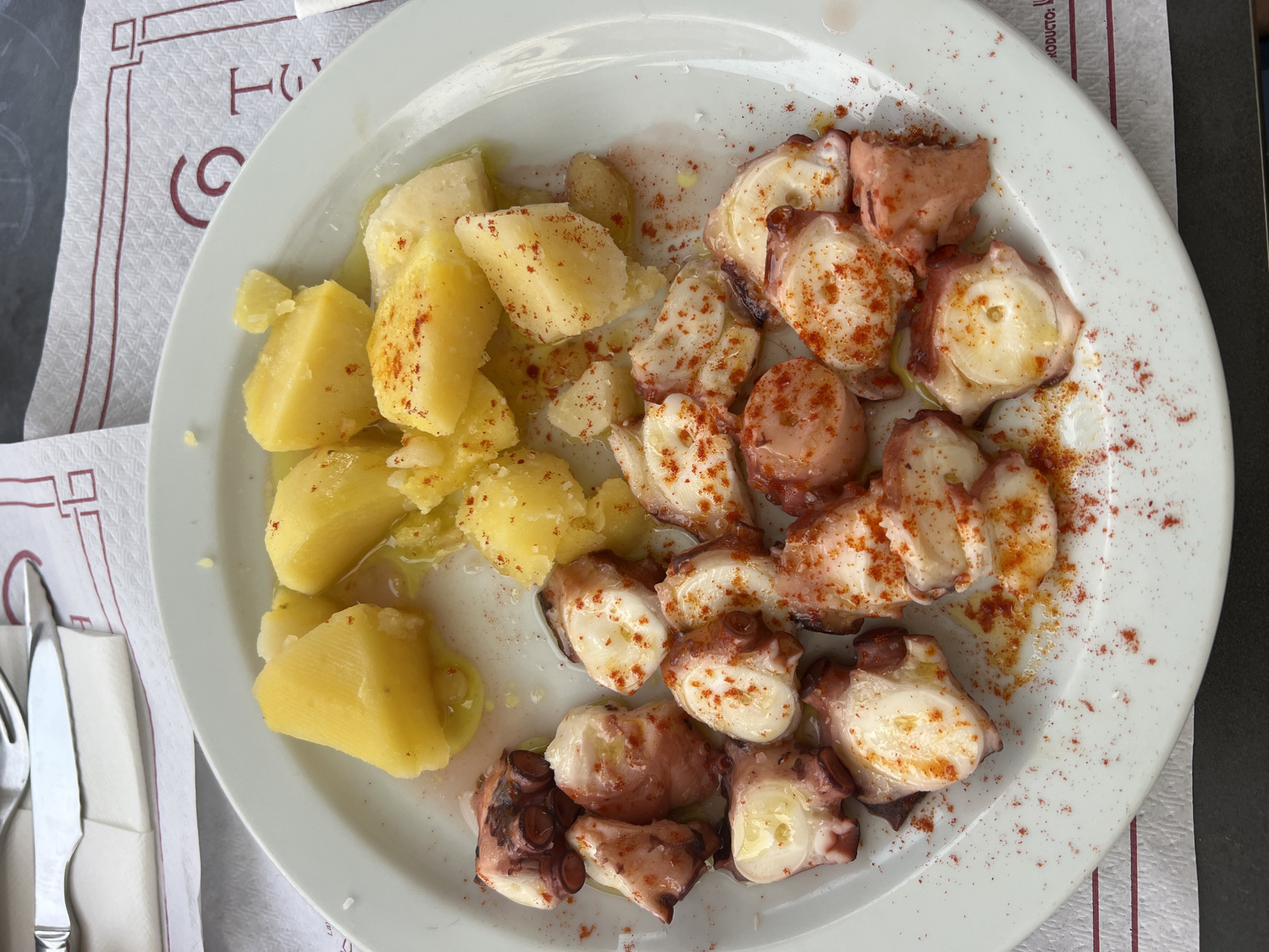 A white plate holds a serving of boiled potatoes and sliced octopus seasoned with paprika. The potatoes are cut into bite-sized pieces and are yellow in color. The octopus is sliced into sections, revealing its white flesh and reddish-purple skin. It is also sprinkled with paprika, some of which has created red speckles on the plate. The plate is on a table covered with a white placemat that has a red border and a stylized "H" and "C" in red lettering. A fork and knife are visible in the upper left corner of the image, resting on a folded white napkin. The lighting suggests an outdoor or well-lit indoor setting. The context provided suggests this scene is from Portomarín, Spain.