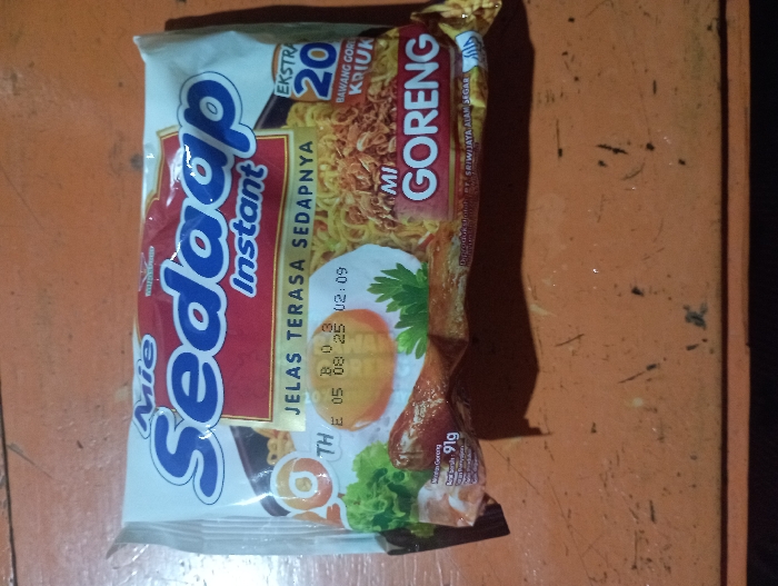 A package of "Mie Sedaap Instant Mi Goreng" instant noodles is centrally positioned in the image, resting on a textured, orange-brown wooden surface. The surface appears worn, with visible scratches and specks, suggesting a rustic or aged setting in Pubian, Indonesia.
The noodle package itself is brightly colored, primarily white with a prominent red and blue logo. The brand name "Mie Sedaap" is displayed in large blue letters, with "Instant" written in smaller script beneath it. The flavor "Mi Goreng" (fried noodles) is clearly visible in red text on a yellow background. The packaging highlights "JELAS TERASA SEDAPNYA," which translates to "Clearly Tastes Delicious." A notable feature mentioned is "EKSTRA 20 BAWANG GORENG KRIUK," indicating extra crispy fried shallots.
An appetizing image of prepared instant noodles with a fried egg, fresh green lettuce, and what appears to be a piece of fried chicken or meat is depicted on the front of the package. The net weight of the product, "91g," is also visible. Below the depicted food, there is text indicating "PT. SRIWIJAYA ALAM SEGAR," likely the manufacturer or distributor.
A timestamp or batch code "B 03 THE 05 08 25 02:09" is printed on the package, which likely indicates a production or expiry date, possibly August 25, 2025, with a time stamp of 02:09 AM/PM. The lighting in the image suggests an indoor setting, with shadows cast by the noodle package, indicating a single light source, possibly artificial. No people or specific activities are visible; the image focuses solely on the product.