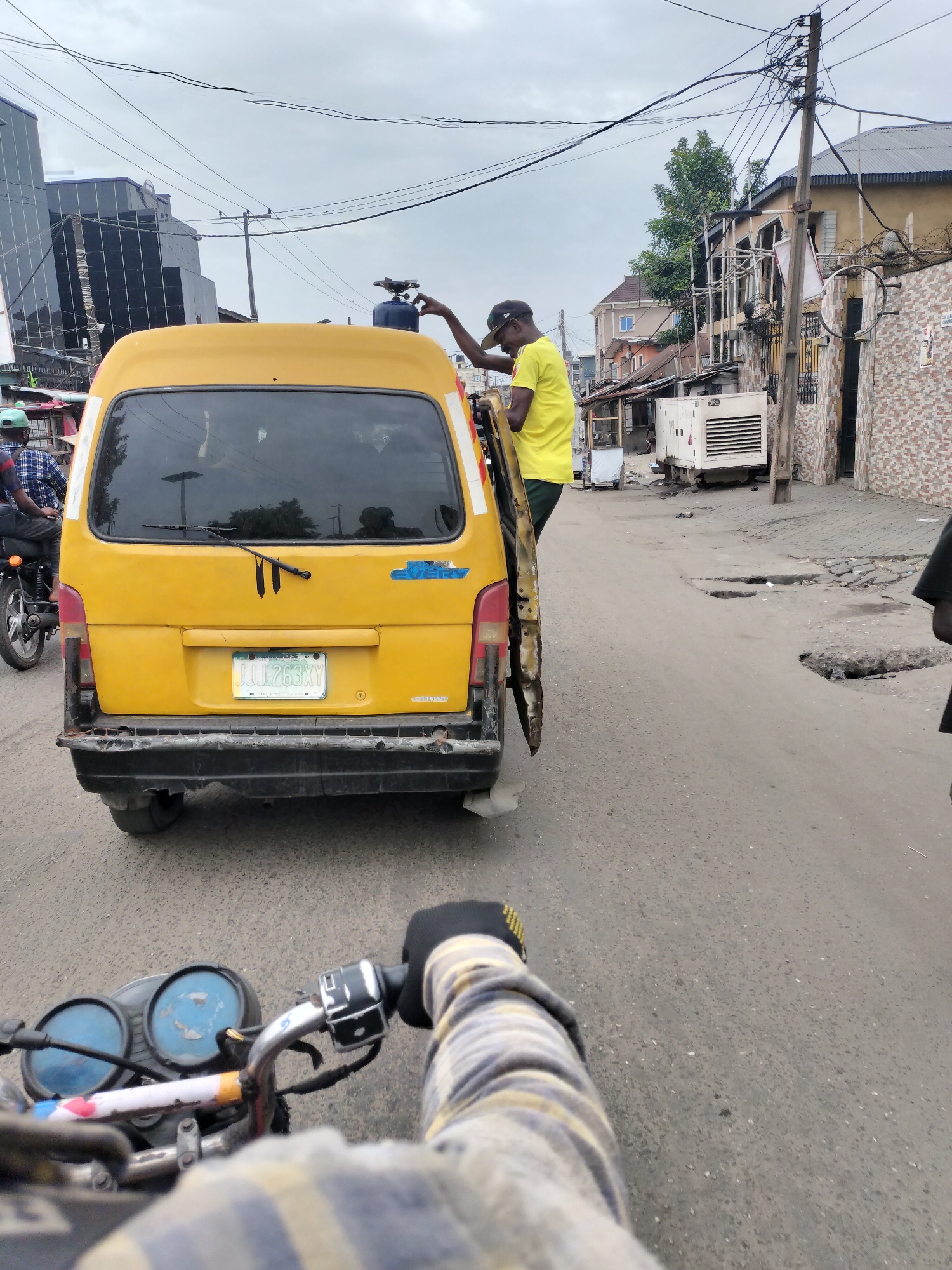 The image depicts a street scene in Alaiyabiagba, Nigeria. A yellow van is positioned in the middle of the road with its back door open. A man wearing a yellow shirt and black cap is standing next to the van, reaching towards a blue object on the van's roof. The van's license plate reads "JJJ 263 XY." The road surface is asphalt with visible potholes. A motorcycle with a rider is visible in the foreground, capturing the scene from the rider's perspective. Additional buildings and infrastructure elements, including power lines and a generator, are in the background, suggesting an urban environment.