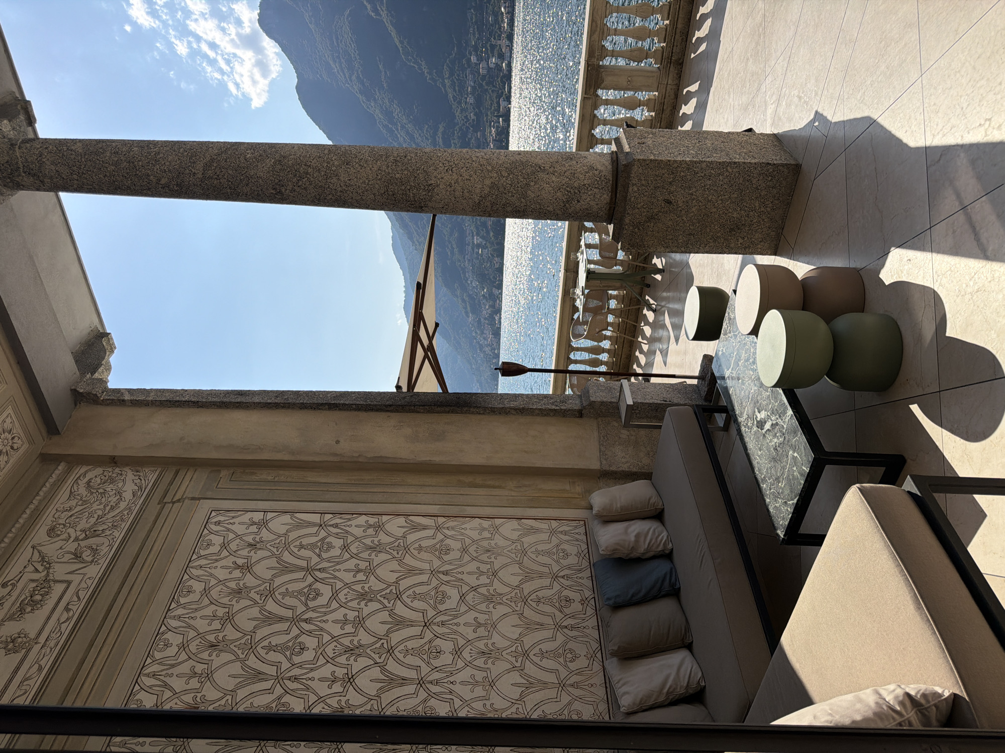 The image shows a shaded outdoor seating area overlooking a body of water and mountains. In the foreground, there is a long, upholstered sofa with several cushions, accompanied by a rectangular coffee table with a marble top and a dark metal frame. Next to the coffee table are three round stools of varying colors: green, beige, and brown. The seating area is on a tiled patio, protected by a stone column and an overhead structure.
Beyond the patio, a white balustrade lines the edge of a terrace. On this terrace, there are small tables and chairs, and a large umbrella provides shade. The water is sparkling under the sunlight, and mountains rise in the background, dotted with buildings. The sky is clear and blue, with a few white clouds. The overall scene suggests a luxurious and serene setting, possibly a hotel or a private residence in Pognana Lario, Italy, given the characteristic lake and mountain landscape.