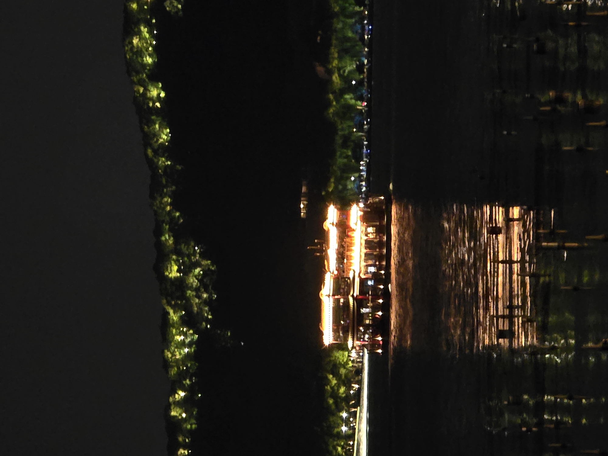 The image captures a nighttime scene on West Lake in Xihu District, China. A multi-decked boat, adorned with extensive warm golden-orange illumination, is prominently positioned on the water, slightly right of the frame's center. The boat's intricate architectural details, including tiered roofs and railings, are highlighted by the artificial lights. Directly below the boat, its lights cast shimmering, elongated reflections on the gently rippled water surface. In the background, a dark silhouette of hills is visible, dotted with numerous small, yellowish-green lights, indicating illuminated trees or features on the landscape. Along the distant shoreline, particularly to the left of the boat, a line of similarly illuminated trees can be observed. The sky above is dark and featureless. No specific individuals are discernible within the frame.