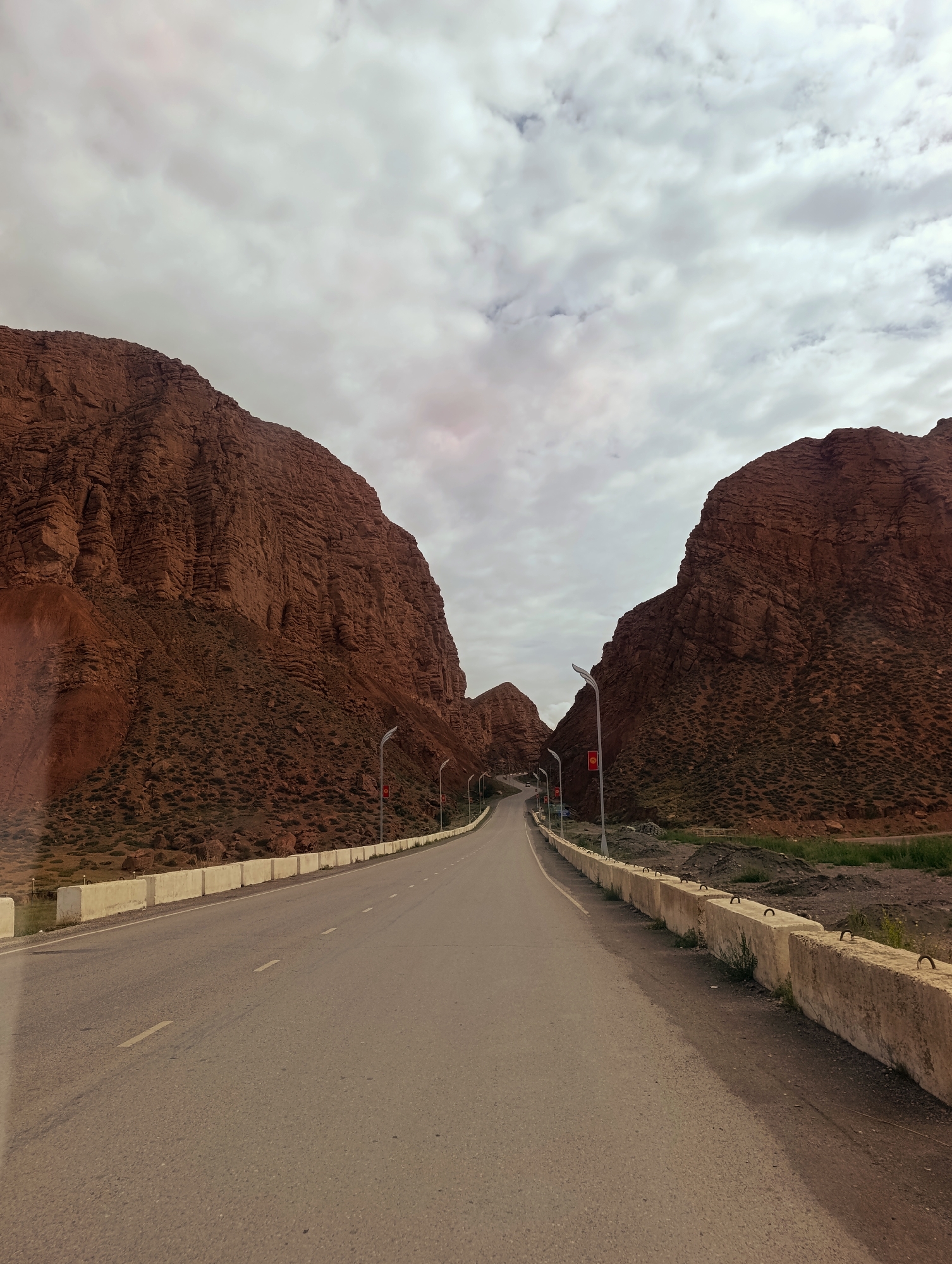 The image depicts a road passing through a gorge with towering, reddish-brown rock formations on either side. The road is paved and appears to be a two-lane highway. There are concrete barriers along the sides of the road. Streetlights with red flags are visible at intervals along the road. The sky is overcast with white and gray clouds. The landscape suggests a mountainous or desert region. The overall impression is of a scenic, remote location, likely a road that is a tourist attraction or a main thoroughfare through a natural wonder. The specific location in the city of Naryn, Kyrgyzstan, is not explicitly identifiable from the visual information alone, but the geology is consistent with the mountainous terrain found in and around Naryn.