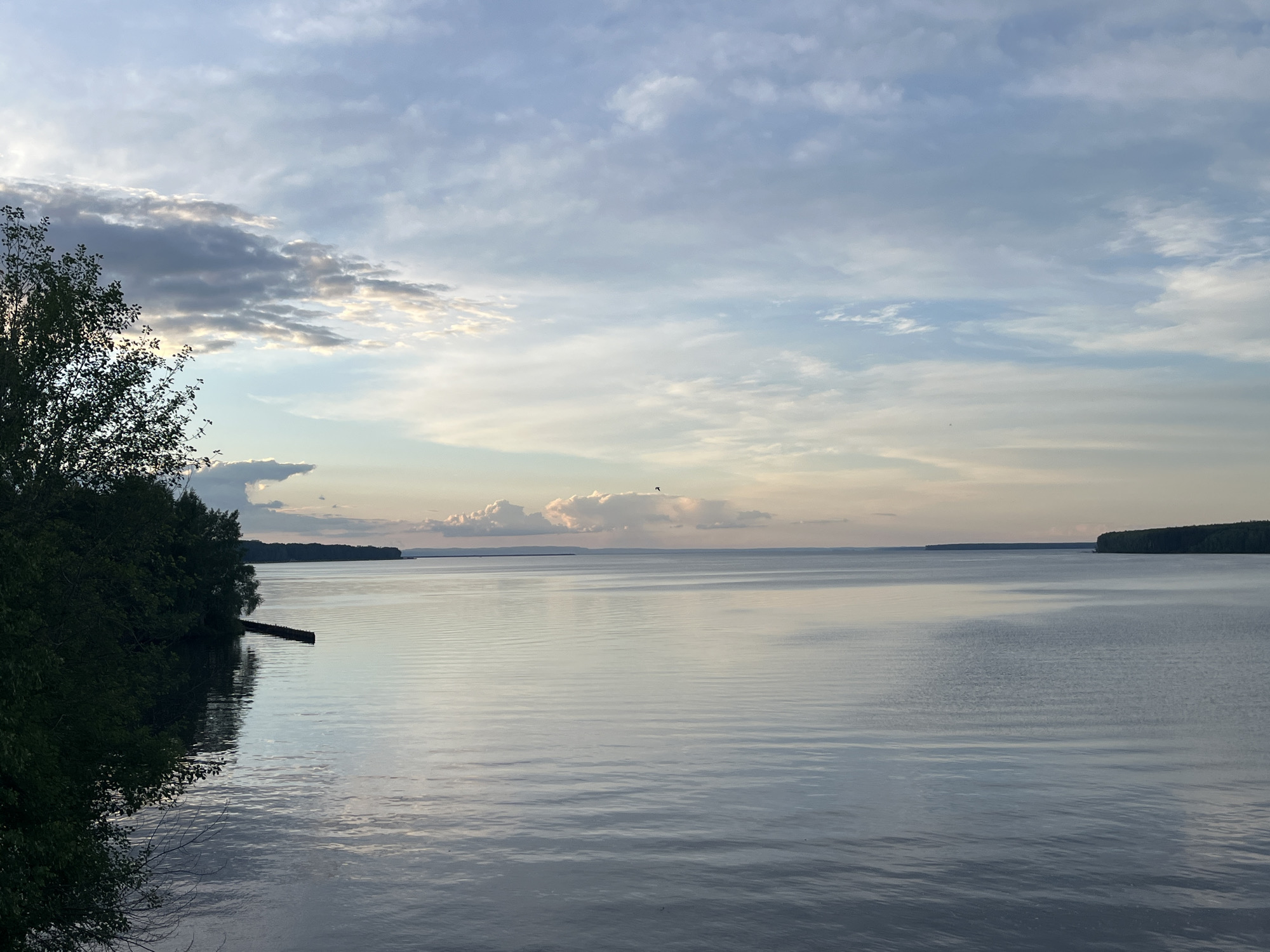 The image depicts a calm body of water, likely the Kama River, in the city of Chaikovsky, Russia. The water surface is mostly smooth with slight ripples reflecting the sky. A line of trees is visible on the left side of the frame, partially obscuring the view, while the horizon is dominated by distant landmasses on either side of the river. The sky is overcast with a mix of grey and light blue clouds, suggesting twilight or early morning. A single bird is visible in the distance over the water. A simple wooden structure protrudes slightly into the water on the left. The foreground is out of focus, giving the impression that the photo was taken from an elevated position.