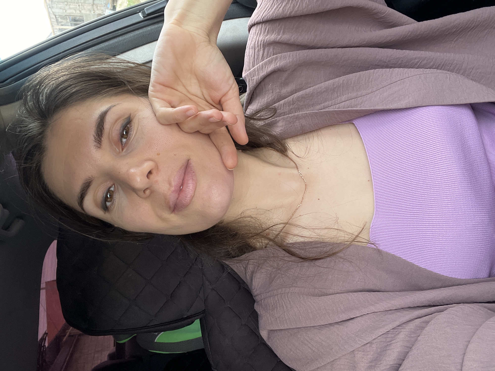 A medium, close-up shot captures a woman within a vehicle. She is resting on a black headrest with green trim. The woman is wearing a purple, ribbed top overlaid with a mauve, crinkled garment. Her hand is raised to her face. The location is within Kaspiysk, Russia.