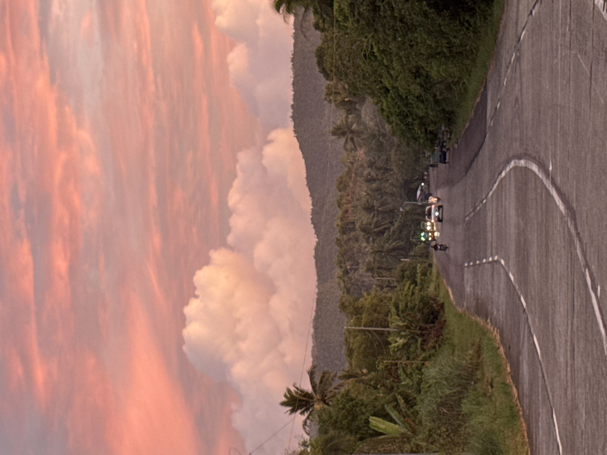The image shows a curved road in Pilar, Philippines, flanked by lush greenery and a mountain range. A pink-tinged sky with large, billowing clouds dominates the left side. Several vehicles, including cars and motorcycles with headlights visible, are traveling on the road. White lines are painted along the road, marking lanes. The scene is likely captured during dusk or dawn, given the sky's color. The location in Pilar is suggested by the typical tropical vegetation and landscape.
