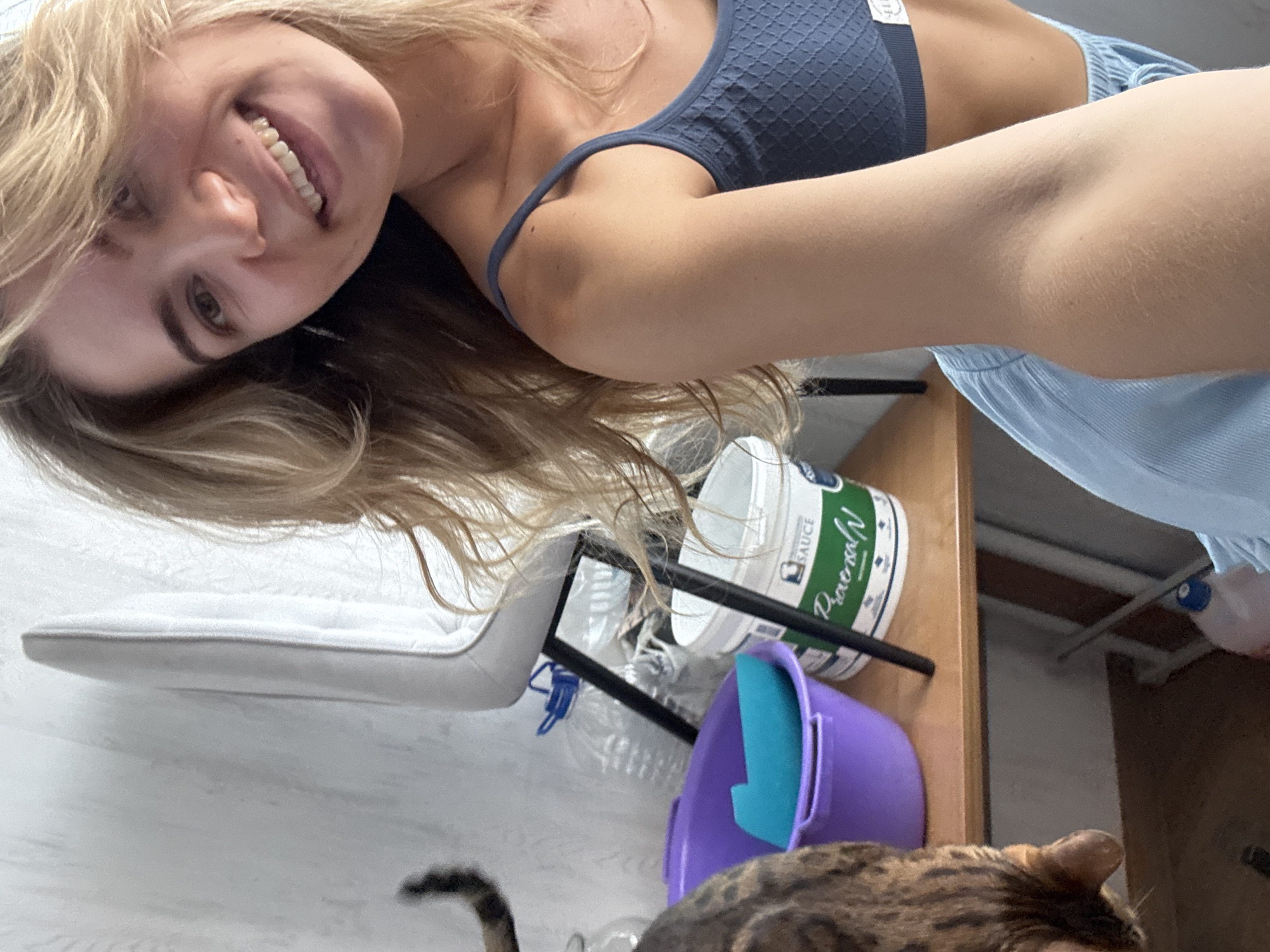 In an indoor setting, a young woman with blonde wavy hair and a bright smile is captured in a selfie. She is wearing a blue textured tank top and light blue shorts. To her left, a cat with tabby markings is partially visible, its tail curved upwards as it leans over a wooden shelf. On the shelf, a purple bowl contains a blue object, and a white bucket with green lettering is also present. In the background, a white chair and plastic bottles are visible.