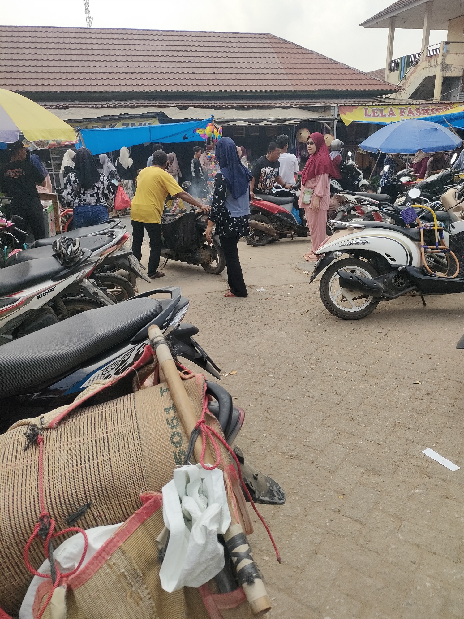 In Desa Banyumas, Indonesia, a bustling outdoor market scene unfolds. A crowd of people, many wearing hijabs, are gathered amidst a collection of parked motorcycles. Several individuals are engaged in transactions or social interactions. A man in a yellow t-shirt appears to be tending to a grill or cooking station, with smoke rising from it. A woman in a pink outfit stands near a stall labeled "LELA FASHION." The ground is paved with interlocking bricks, and several large, woven sacks are visible in the foreground, secured with ropes. Umbrellas, both yellow and blue, provide shade. The architecture in the background suggests a mix of single-story buildings with tiled roofs and a multi-story structure. The overall atmosphere is that of a lively community gathering.