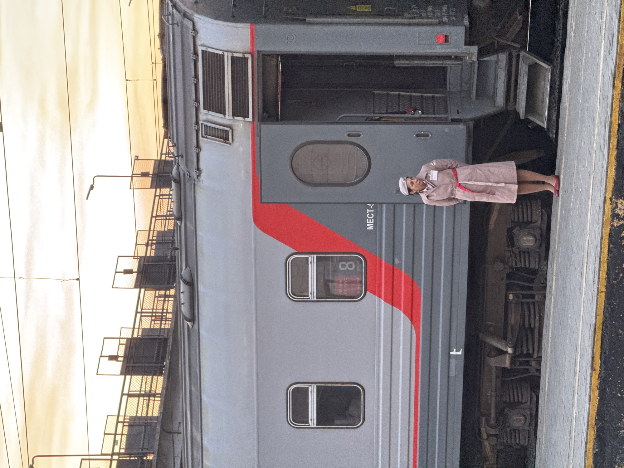 A uniformed woman stands on a station platform next to a train. The train is gray with a red stripe. The woman is wearing a light pink coat, a white hat, and red shoes. The sky is golden in the background. Based on the text visible on the train's window, the location is Izhevsk, Russia.