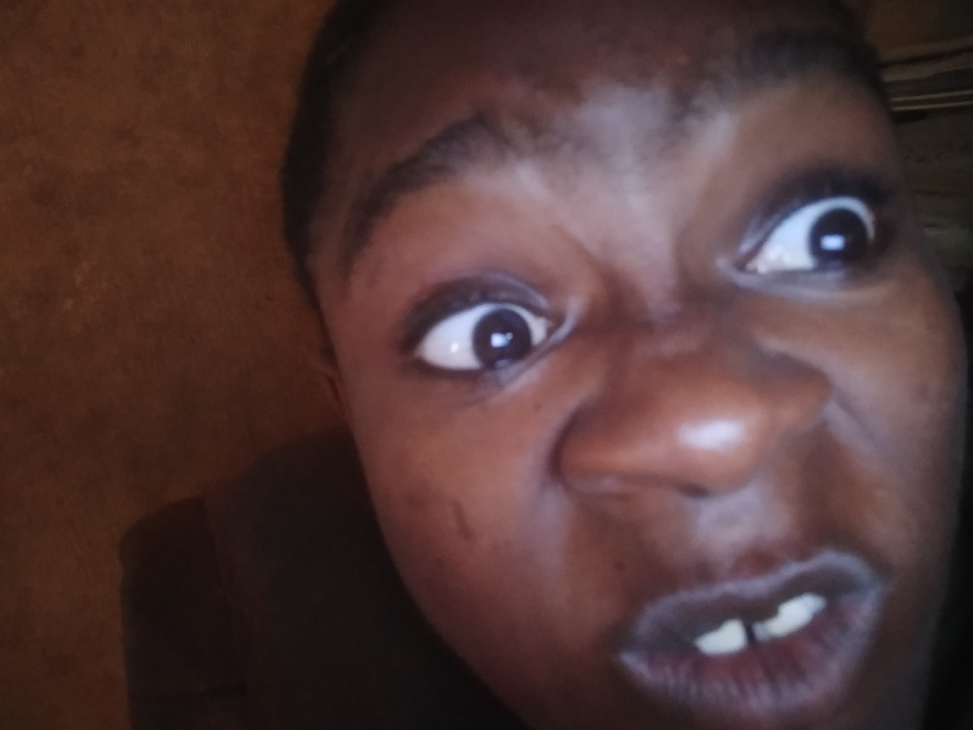 Close-up of a person's face. The person has brown skin, dark eyes, and short dark hair. Their eyebrows are furrowed and their eyes are wide open, with the pupils dilated. The nose is prominent. The mouth is open slightly, revealing teeth. The background is a blurry brown color. The photo shows a close-up perspective, focusing on the person's facial expression.
I cannot give a precise location, as none is clearly provided.