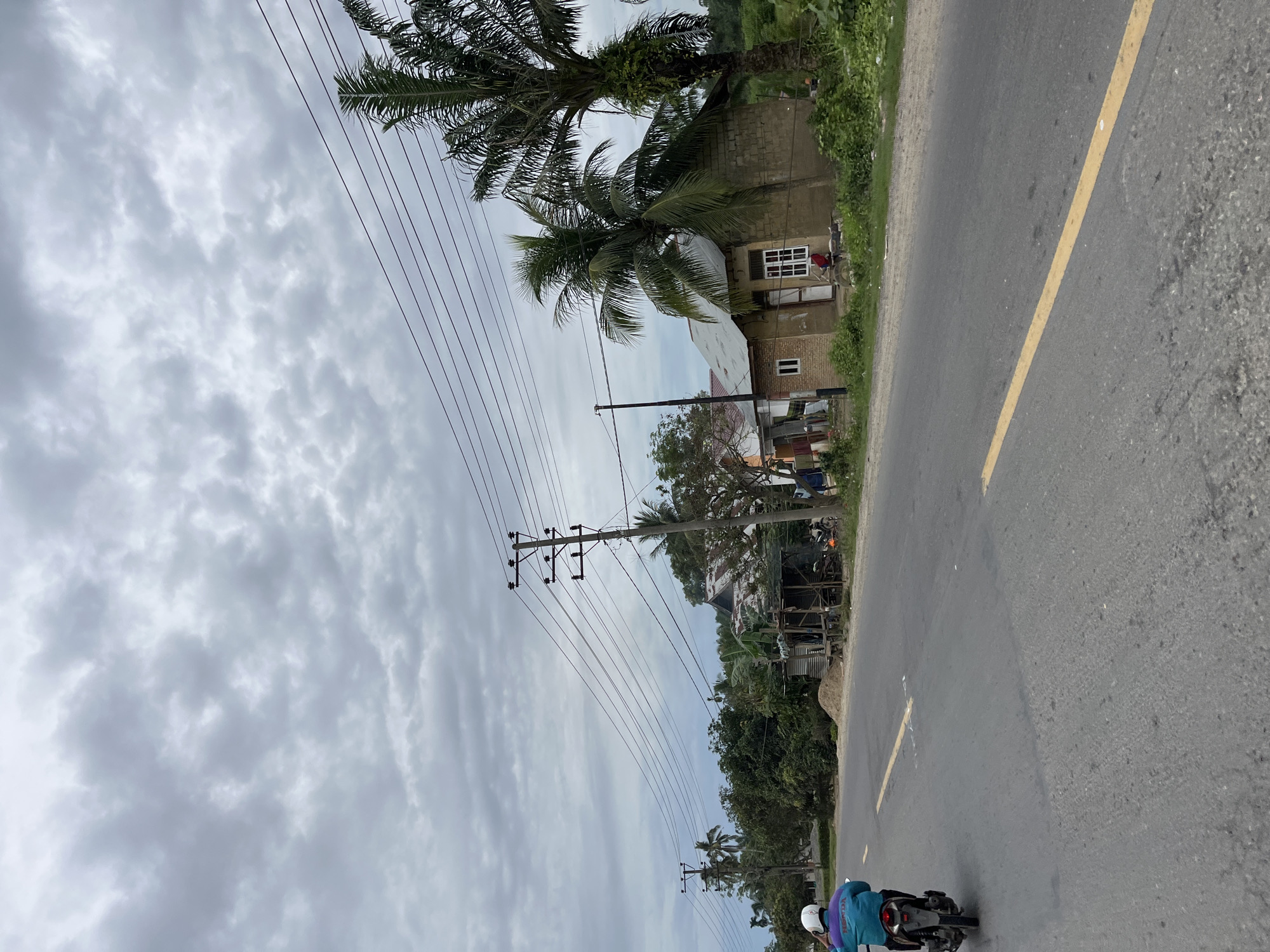 The image depicts a street scene in Simpangempat, Indonesia, under a cloudy sky. A paved road with a yellow dividing line runs diagonally across the frame. On the right side of the road, there's a small, light-colored building with multiple windows. Palm trees, with their distinctive fronds, are visible in front of and to the side of the building. Power lines crisscross the sky, supported by utility poles.
Further down the road, more vegetation and smaller structures can be seen. In the foreground, a motorcycle with two occupants is traveling on the road. The person in the back is wearing a blue jacket and a white helmet, while the person in the front is wearing a white helmet. The overall impression is of a typical day in a semi-urban or rural area in Indonesia, with everyday life unfolding along the roadside. The weather appears overcast, suggesting it might be daytime, possibly late morning or afternoon, given the overall brightness of the scene. There are no visible signs with text in this image.