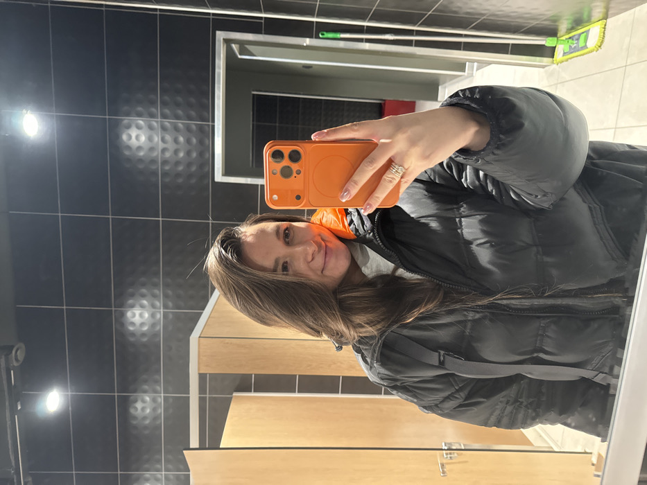 A woman wearing a black puffer jacket and an orange inner layer poses for a selfie in a bathroom. She is holding an orange smartphone in her right hand. The bathroom features black tiled walls with a textured pattern and light-colored wooden doors. A mop leans against the wall behind her, and the reflection of the scene is visible in the mirror.