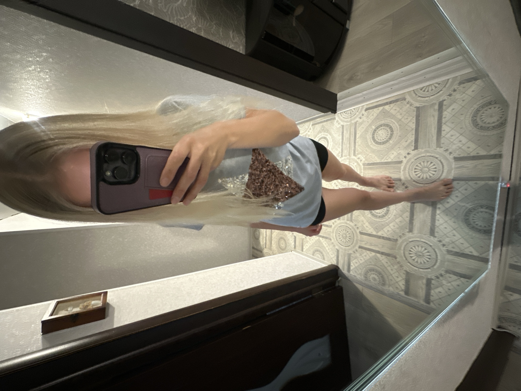 The image is a mirror selfie taken indoors, likely in a hallway or entryway. A young person with long blonde hair is visible, holding a smartphone to take the picture. They are wearing a light grey t-shirt with a sequined star design on the front and black shorts. The background shows a dark wooden door on the left, a wall with a small framed picture, and a patterned floor. On the right side, a dark cabinet is visible, and part of a mirrored surface reflects the scene. The floor tiles have a geometric pattern with circular motifs. The overall environment suggests a residential setting within the city of Energetik, Russia, as specified by the prompt, though no specific landmarks are identifiable.