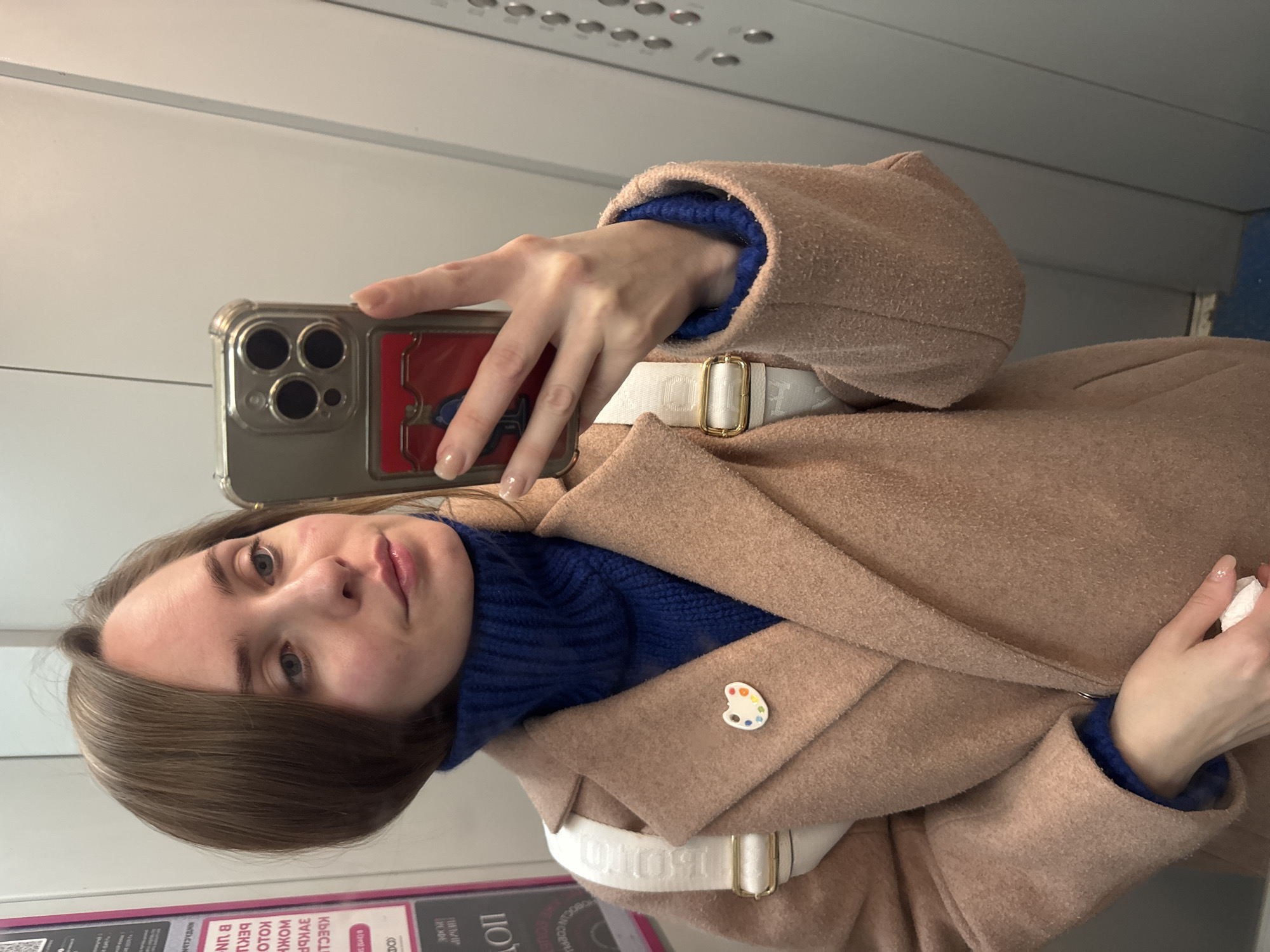 A woman is taking a selfie indoors, possibly in an elevator. She is wearing a tan coat over a blue turtleneck sweater. A white belt is visible over the coat. A small pin shaped like a painter's palette is on the lapel. She is holding a smartphone in her right hand, with a phone case with a red and blue design. The background features elevator buttons and interior panels. A poster in Russian is visible at the bottom left. The city of Perm, Russia, is mentioned on the poster at the bottom left corner of the image.