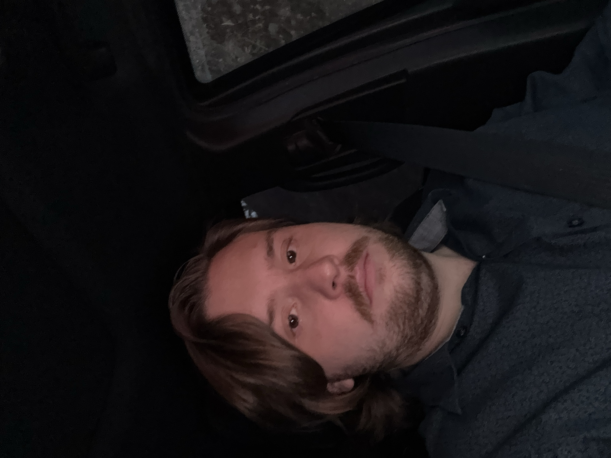 A man is seated in a car, wearing a blue button-down shirt and a seatbelt. The environment appears dimly lit, and the focus is on the man's face, which features light brown hair and a beard. The location is within a car, presumably in transit. There is a window at the top of the frame, suggesting the position of the passenger is in the car, likely in Huixquilucan de Degollado, Mexico. No specific landmarks are visible to confirm the precise location.
