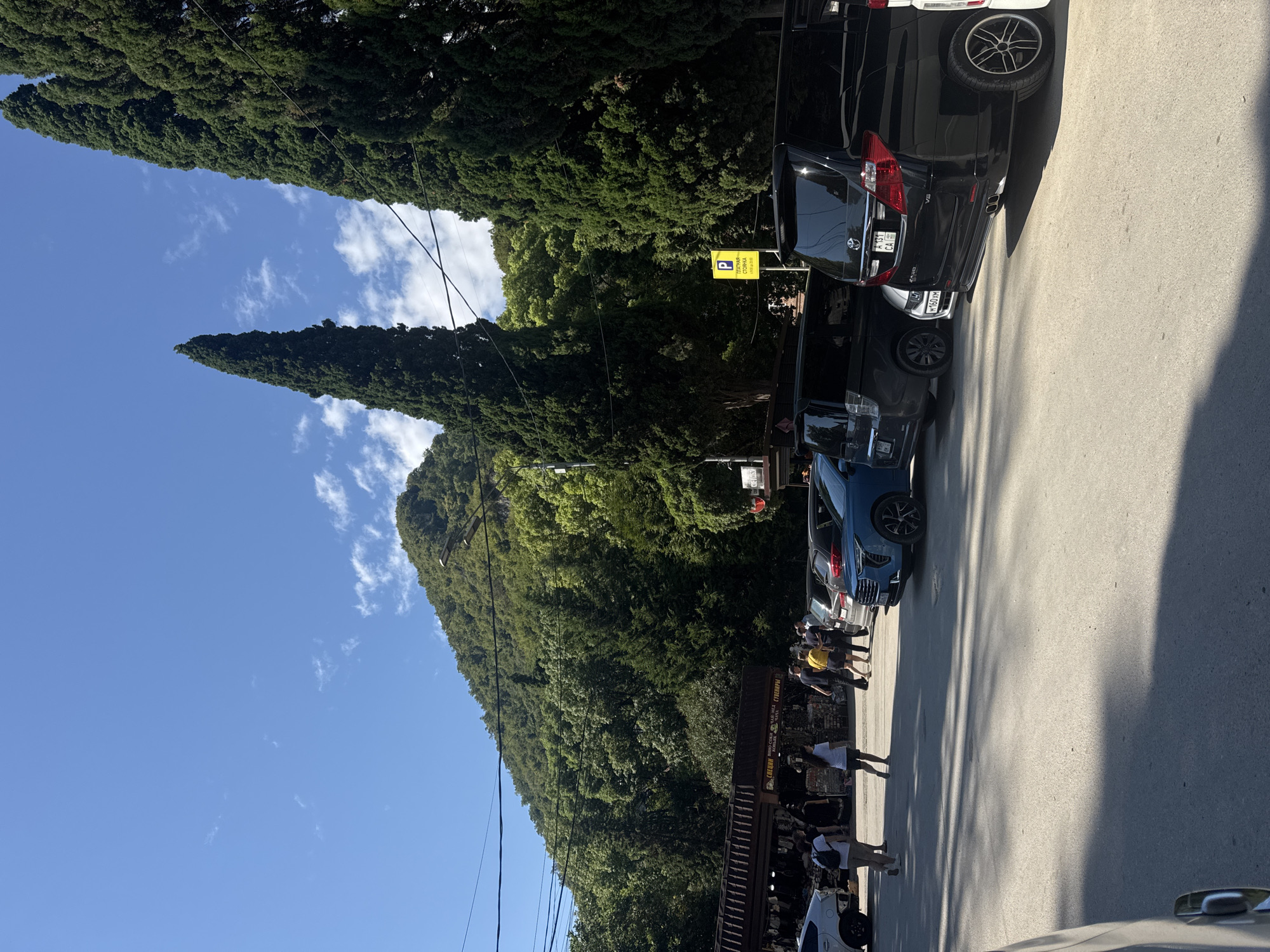 The image captures a scene in New Athos, Abkhazia, under a clear blue sky with scattered white clouds. The background features a lush, green, mountainous landscape. In the foreground, a paved parking area is occupied by several vehicles, including black minivans, a blue SUV, and a silver sedan. A prominent yellow parking sign with the letter "P" is visible. To the left, a row of market stalls with awnings displays goods. Several people are walking in the parking area and near the market. Two tall, dark green cypress trees stand to the right of the frame, with power lines running between them and the mountain. The overall impression is a sunlit day in a tourist or market area situated at the base of a verdant mountain.