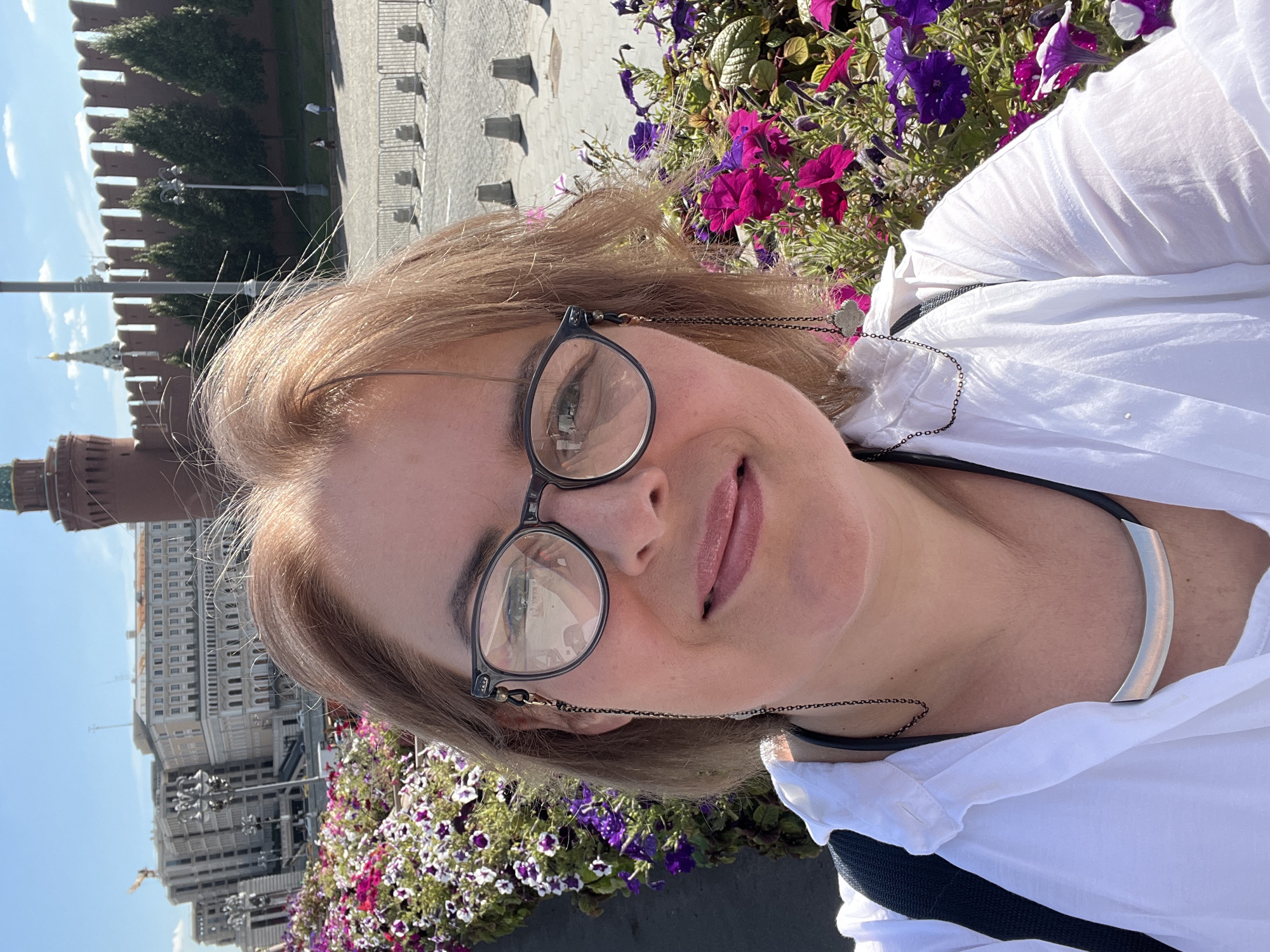 A woman with shoulder-length blonde hair and glasses takes a selfie outdoors with a city skyline in the background. She is wearing a white button-down shirt and a silver necklace. The background includes red brick walls, trees, and buildings, suggesting a historic city location. The flowers in the foreground are a mix of pink and purple petunias. The woman is smiling slightly, and her face is in focus. The lighting suggests it is a bright, sunny day. The architecture and red brick walls are characteristic of the Moscow Kremlin.
