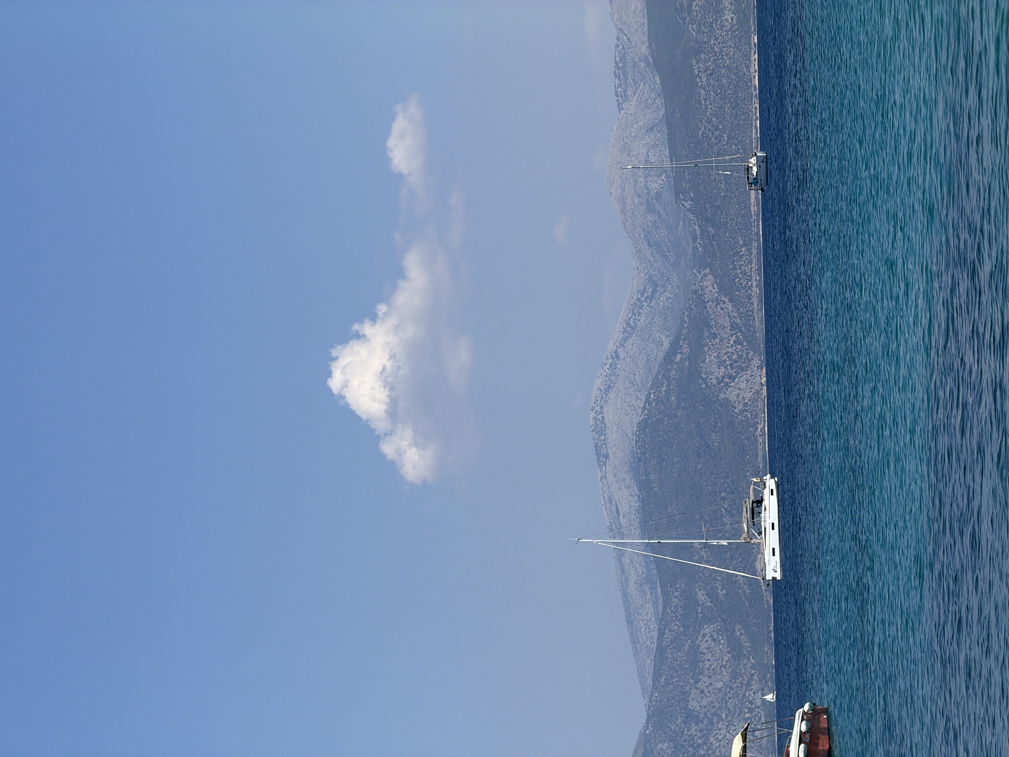 A vertical, medium shot captures a serene coastal scene under a clear blue sky with a solitary, puffy white cloud. The foreground is dominated by deep blue, rippling water, transitioning to a lighter turquoise hue closer to the horizon. Two sailboats are visible in the middle ground; the larger one, white with a tall mast, is positioned to the left of center, sailing with its sails furled. To the right, a smaller sailboat is also visible. In the background, a chain of rugged, gray mountains stretches across the horizon, their slopes textured with sparse vegetation. The sea appears calm, with gentle waves suggesting a peaceful day. The image evokes a sense of tranquility and the vastness of the natural environment. The location is the Municipal Unit of Kastos, Greece, known for its clear waters and scenic islands. There are no people visible in this image.