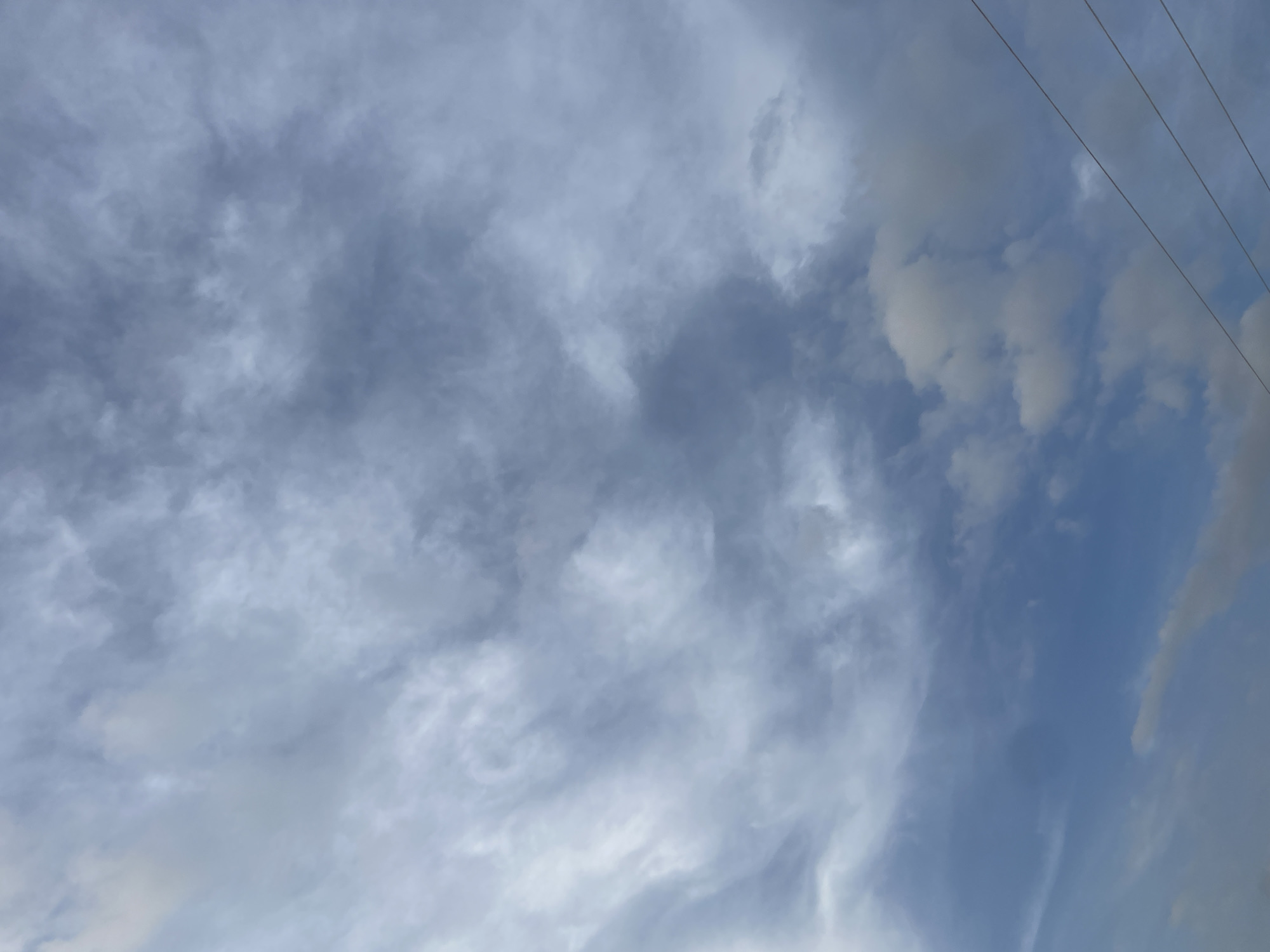 The image shows a cloudy sky above Jalingo, Nigeria. Three thin, dark power lines are visible in the lower right corner, running diagonally upwards. The clouds are a mix of gray and white, with some areas appearing darker and more dense. The sky is a pale blue in the lower portion of the image. There are no people or structures visible in the scene.