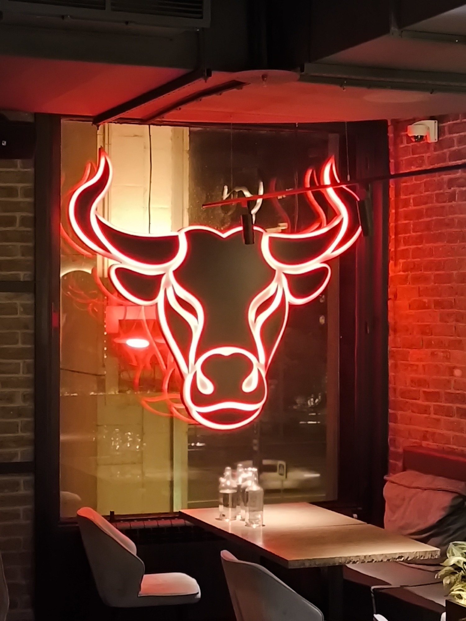 A close-up shot captures a vibrant red neon sign shaped like a bull's head, centrally positioned against a window in what appears to be a restaurant or bar. The bull's head, outlined in glowing red, features prominent horns and a stylized face. Behind the neon sign, the window reveals a blurred nighttime city scene, possibly in Moscow, Russia, with streetlights and reflections of interior lights. To the right, a red brick wall is visible, adding to the establishment's ambiance. In the foreground, a table is set with bottles of water and a few chairs, one of which is visible on the left, upholstered in a light gray fabric. The lighting is predominantly red, casting a warm and inviting glow across the scene.
