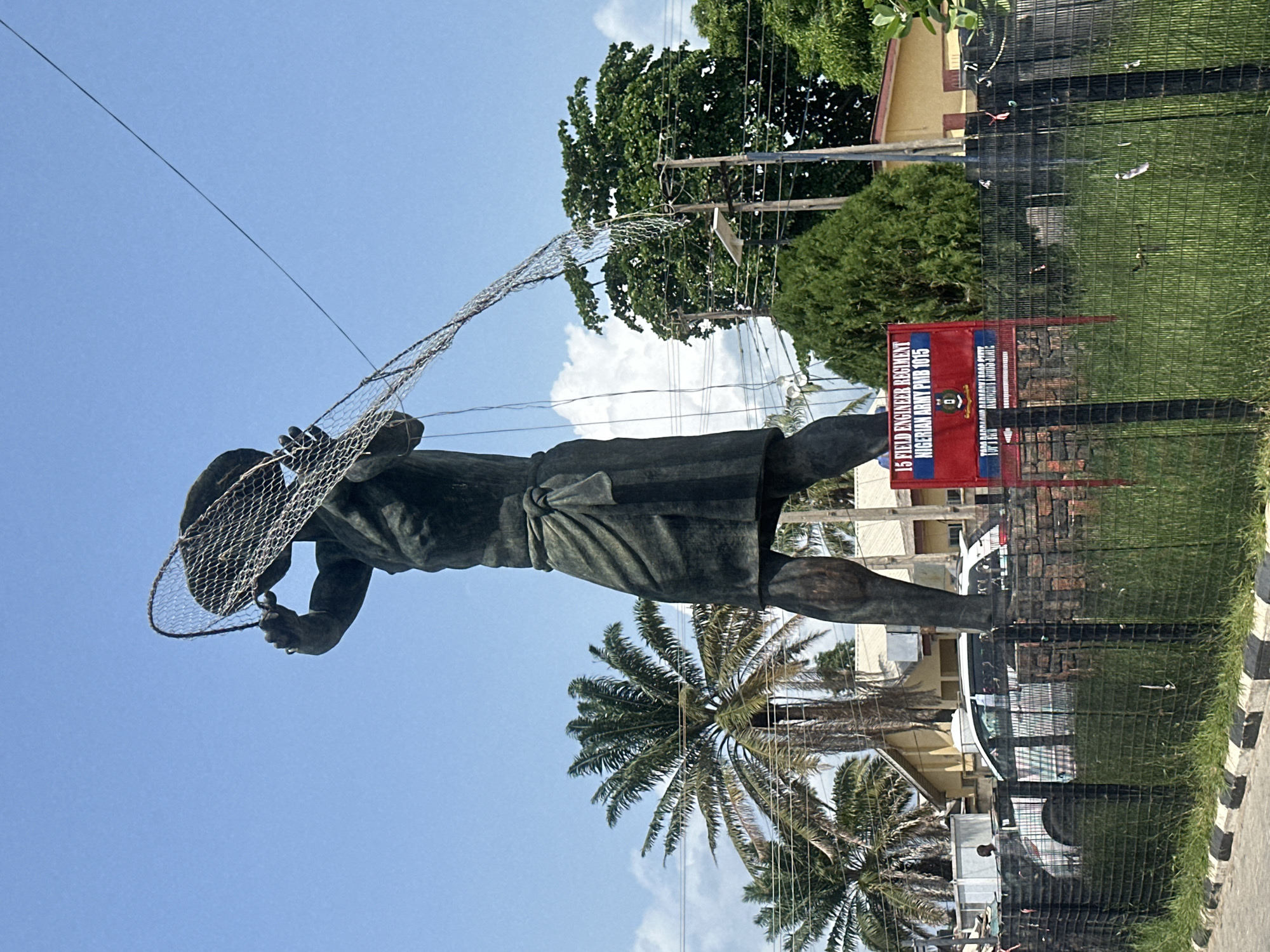 This image, taken in Badagry, Nigeria, features a large, dark-colored statue of a fisherman actively casting a net, positioned prominently on the left side of the frame. The statue depicts the man with a determined posture, holding the wide fishing net with both hands as if mid-throw. He wears a simple wrap around his waist.
The scene is set outdoors under a bright, clear blue sky with a few sparse white clouds, indicating sunny weather during the day, likely midday or early afternoon. Numerous power lines crisscross the upper and middle sections of the background, supported by utility poles. Lush green foliage from various trees, including several tall palm trees, fills the background, suggesting a tropical environment.
Behind the statue and across the mid-ground, a chain-link fence runs horizontally, separating the immediate foreground from further background elements. Beyond the fence, green grass is visible, followed by several buildings with light-colored walls and reddish-brown roofs. A red rectangular sign stands out against the greenery behind the fence. The text on this sign clearly reads: "15 FIELD ENGINEER REGIMENT" and "NIGERIAN ARMY PUB 1015". Below this, smaller text indicates "TOP CANOPY ACADEMY BADAGRY". A black and white striped curb or barrier is visible towards the bottom right, suggesting the edge of a road or pathway.