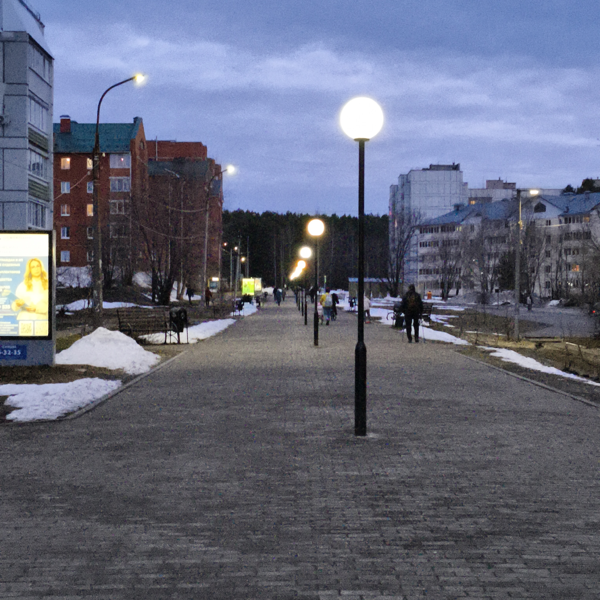 The image captures a twilight scene on a city walkway in an unidentified location. The walkway, paved with gray bricks, stretches into the distance where people are walking. It is lined with evenly spaced, spherical streetlights that illuminate the path. On either side of the walkway are patches of melting snow, indicating it is late winter or early spring.
Buildings of varying heights and construction styles are visible on the sides of the walkway. One building is brick with a teal-colored roof, while others are gray and appear to be apartment complexes. A billboard featuring a woman is located on the left side of the walkway. The sky is overcast with muted blue and gray tones. Some trees stand bare in the background, between the buildings. Some people are walking alone while others are in small groups. A few benches are placed along the walkway.