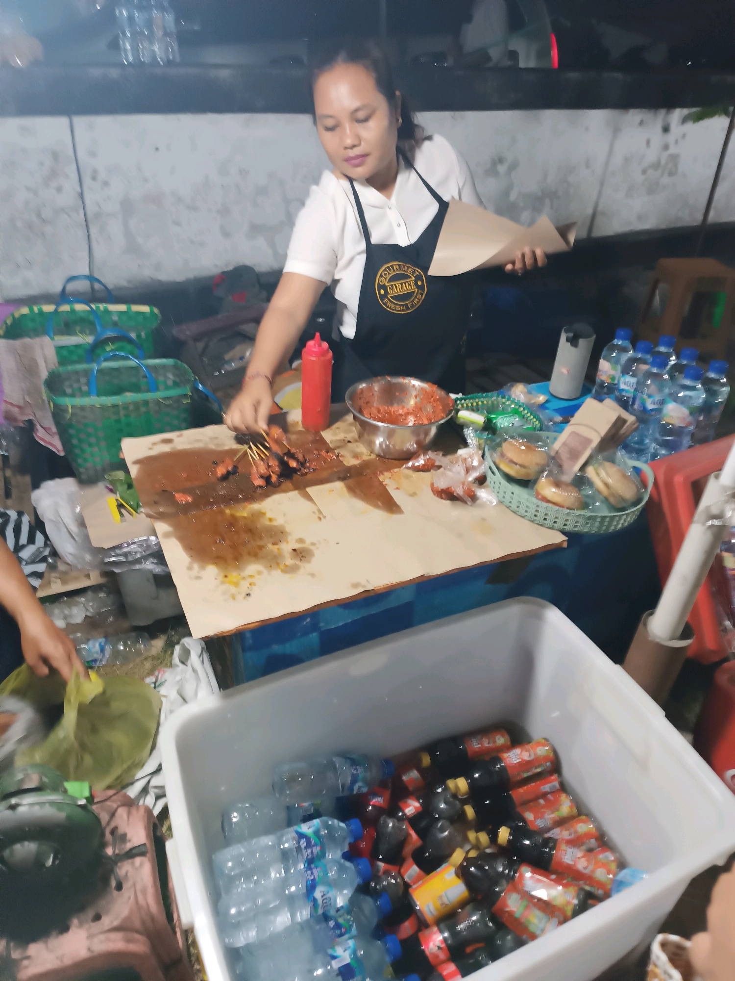 A woman stands behind a food stall in Kediri, Indonesia, preparing food. She is wearing a white shirt and a black apron with a logo that reads "Gourmet Garage Fresh First". She is holding a piece of brown paper in her hands. In front of her is a wooden board with satay skewers, a bowl of chili sauce, a red squeeze bottle, and several burgers in plastic wrap. To her left are two green woven baskets. In the foreground, a white cooler is filled with bottles of water and soda. The stall appears to be set up outdoors, possibly on a street or market.