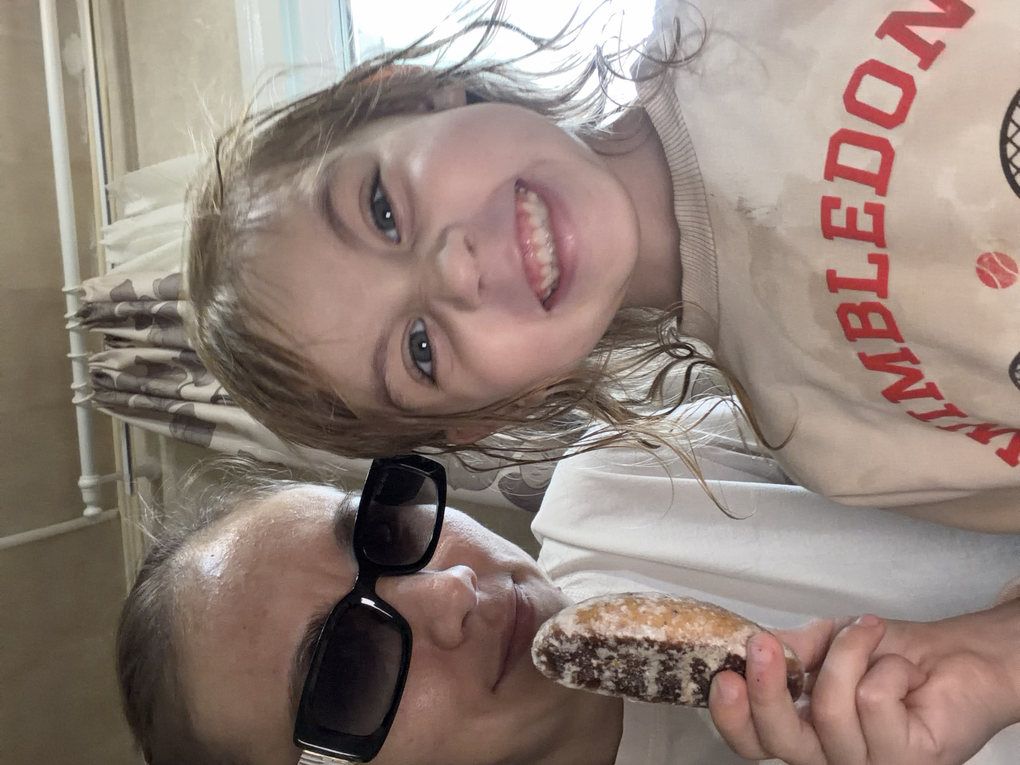 Two individuals, an adult and a child, are posing for a selfie. The adult, on the left, is wearing sunglasses and a white t-shirt, and is holding a pastry in their left hand. The child, on the right, is wearing a light-colored sweatshirt with red lettering that appears to say "WIMBLEDON". The background consists of a curtain and a wall. The lighting suggests an indoor setting. The image appears to have been taken in a residential environment in Tambov, Russia.