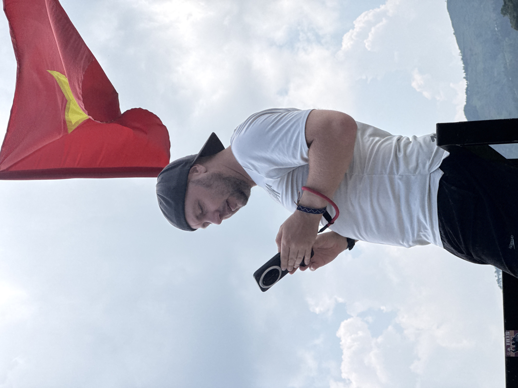 A person stands outdoors in Lũng Phìn Commune, Vietnam, holding a black electronic device with a circular detail. They wear a white t-shirt, black bottoms, a dark cap, and wrist accessories. A red flag with a yellow star is prominent to the left, and a cloudy sky fills much of the background. Part of a mountain or hill is visible in the upper right.