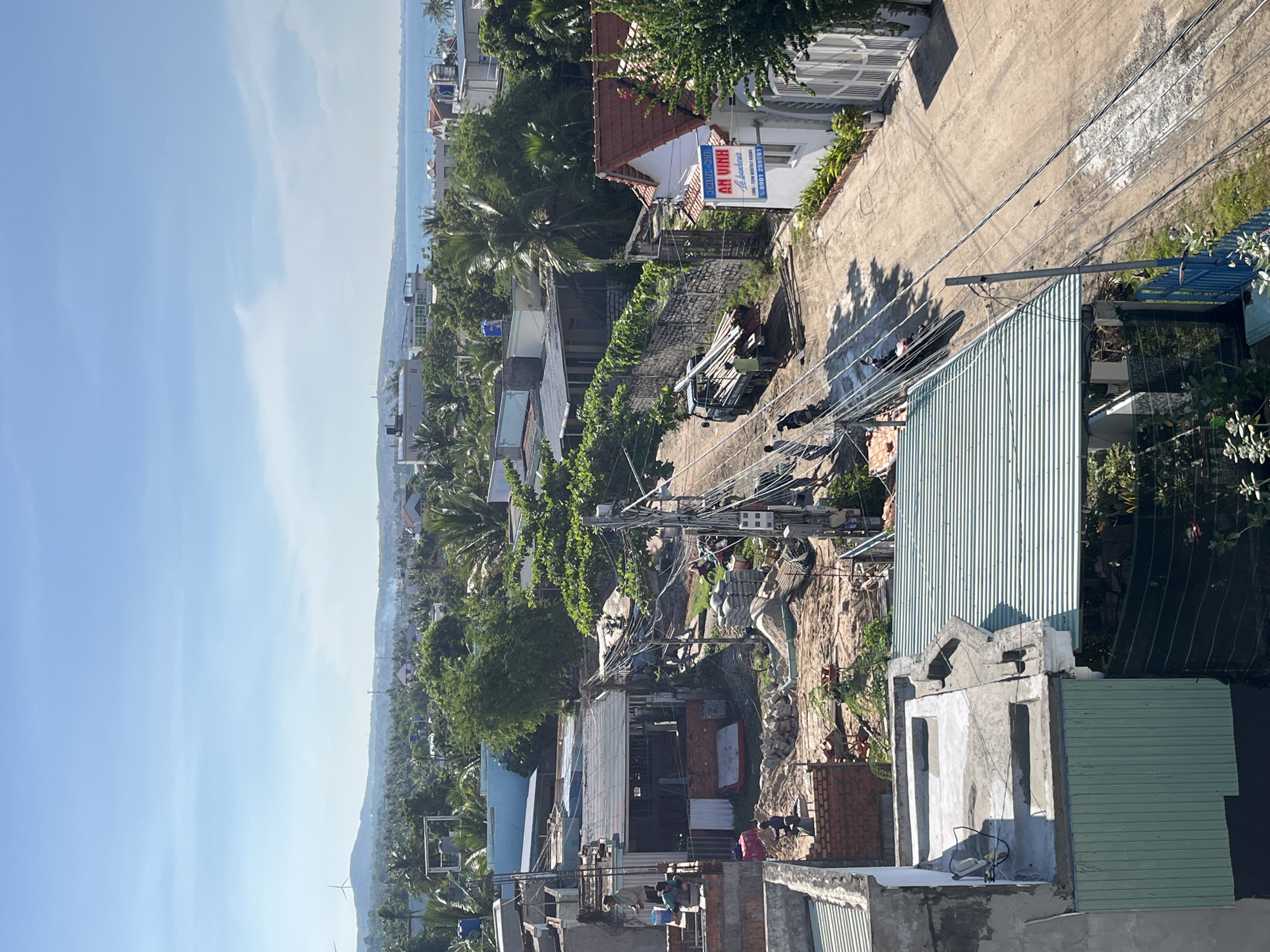 This is a high-angle, medium-shot image taken in Mũi Né, Vietnam, under a clear blue sky with wispy clouds. The foreground features rooftops, some with corrugated metal in shades of green and grey, and others with exposed concrete. A construction site is visible, with piles of sand, bricks, and cement bags, as well as workers engaged in building activities. Electrical wires crisscross the scene, supported by utility poles.
The middle ground shows a dusty road with a small truck carrying long pipes and a motorcycle. Several buildings, including houses and what appears to be a homestay or hotel with a red-tiled roof and a sign that reads "AN VINH HOMESTAY HOTEL," line the street. Lush green trees, including palm trees, are scattered throughout the urban landscape.
In the distance, a town stretches towards the ocean. The horizon is marked by a range of hills with a wind turbine visible on one of them, and further along, the blue expanse of the sea meets the sky. The overall impression is one of a coastal town undergoing development, with a mix of residential, commercial, and construction activities set against a tropical backdrop.