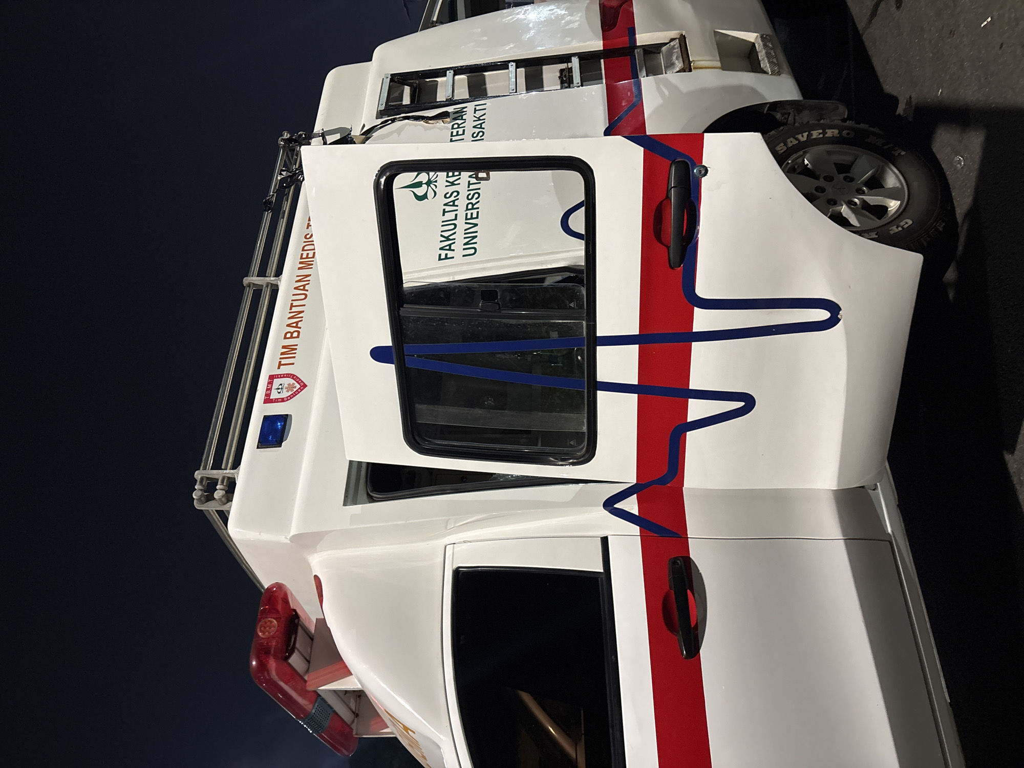 The image shows a white ambulance parked outdoors at night. The ambulance has a red and blue light bar on its roof, a red stripe along its side, and a blue electrocardiogram (ECG) graphic design. Text on the ambulance reads "TIM BANTUAN MEDIS" (Medical Assistance Team) and "FAKULTAS KEDOKTERAN UNIVERSITAS TRISAKTI" (Faculty of Medicine, Trisakti University). The driver's side door is open, and a ladder is mounted on the rear side. The background is dark, indicating it is nighttime. The location is identified as Central Jakarta, Indonesia.