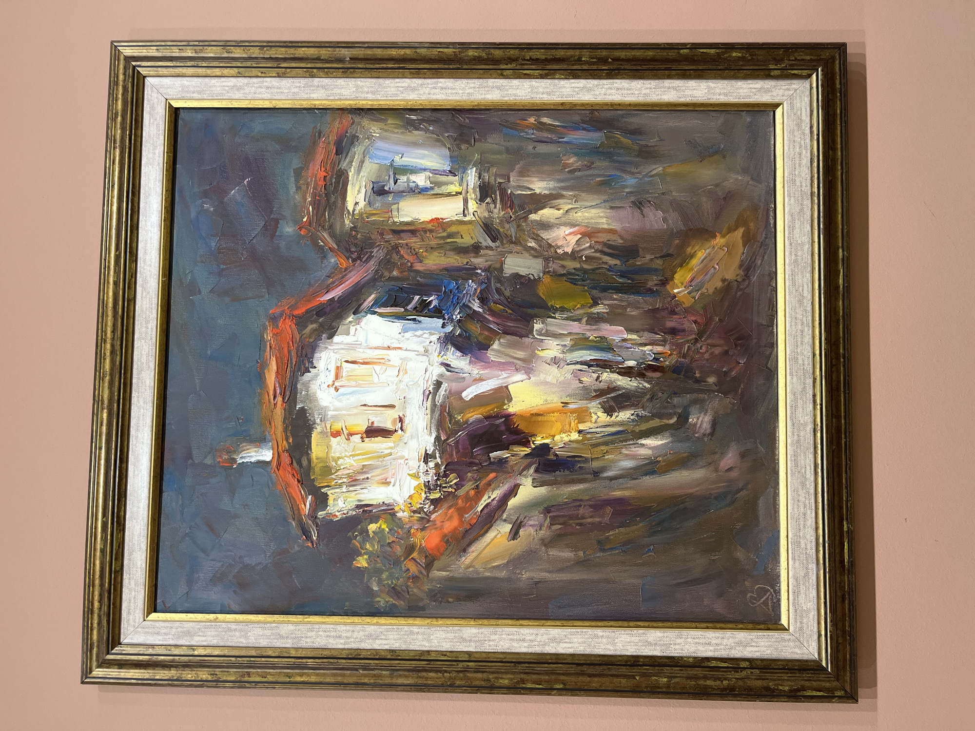 A framed abstract oil painting hangs on a light pink wall. The painting is dominated by gestural brushstrokes in a palette of blues, browns, yellows, and whites. A possible suggestion of a structure or object is discernible in the center, composed of bright white and yellow hues, contrasted against a darker, more atmospheric background. The painting is framed in a multi-layered frame consisting of a gold outer layer, a white intermediary, and a gold inner layer. The lower-right corner of the painting features a signature. The location in Plovdiv, Bulgaria, cannot be determined from the visual information available.