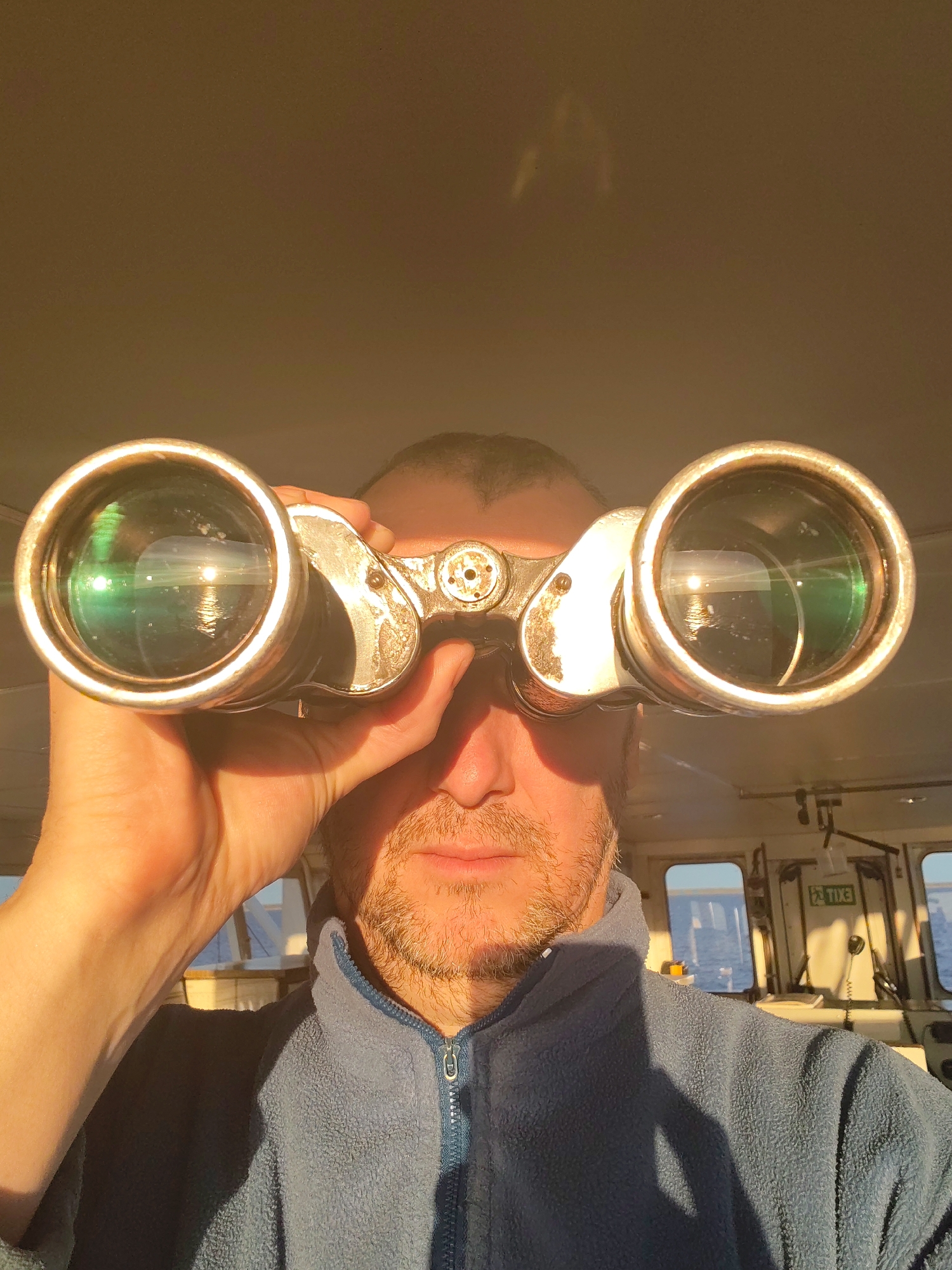 A man is holding binoculars up to his eyes. The lenses reflect the ocean and sky. He is wearing a blue fleece jacket. The background suggests he is inside the bridge of a ship, with windows showing the ocean and an "EXIT" sign visible. The location, Myaksa, Russia, cannot be confirmed from the visual information provided.