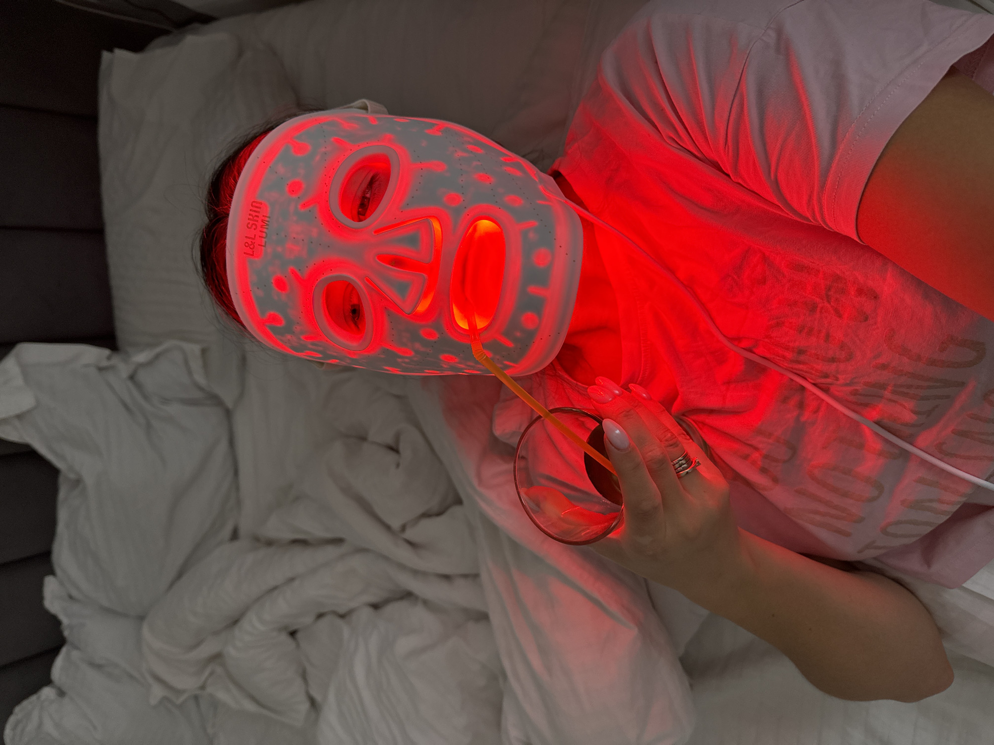 A person, likely female, is depicted from an overhead angle, lying down indoors, presumably in a bedroom in Odintsovo, Russia. They are engaged in a self-care routine, wearing a white or translucent LED light therapy facial mask that emits a prominent red light. This red light strongly illuminates the person's face, neck, and the upper part of their light pink t-shirt, casting a warm glow across the white, crumpled bedding and pillows.
The mask features multiple circular and elongated openings for the eyes, nose, and mouth, through which the red light shines intensely. The words "L&L SKIN LUMI" are visibly printed on the right side of the mask. The person's dark hair is visible at the top edge of the mask.
In their left hand, adorned with a ring, they hold a clear glass containing a dark liquid, possibly a beverage, from which a thin yellow-orange straw protrudes. The overall ambiance suggests a moment of relaxation and pampering in a dim or dark room, where the LED mask is the primary light source.