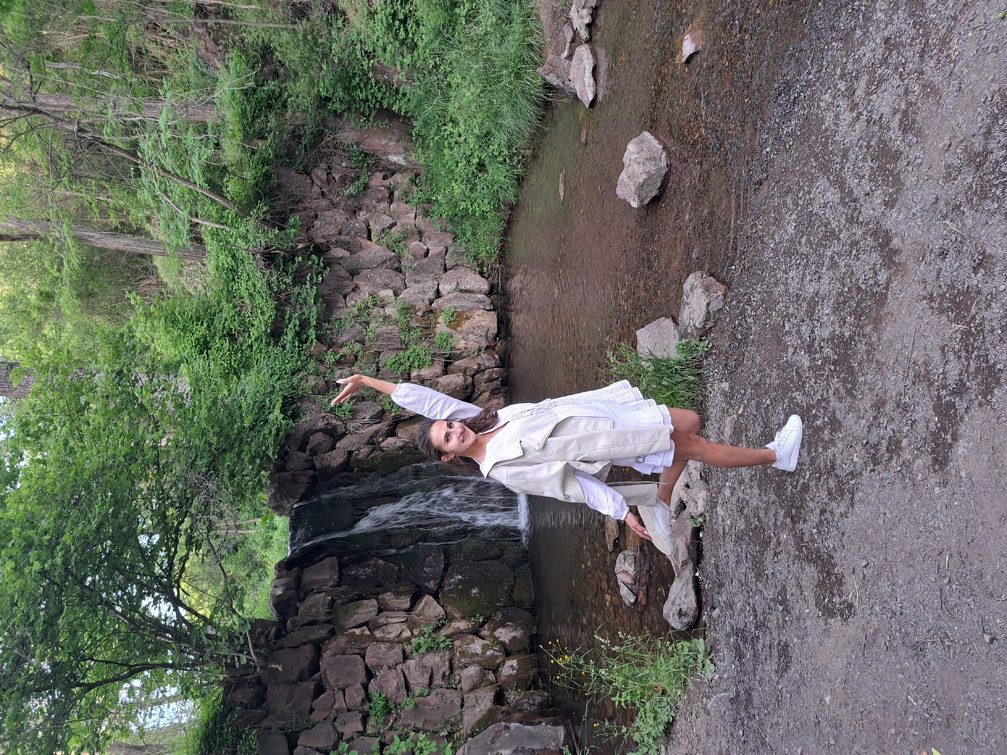 A young woman with dark hair is posing in a natural setting near a small waterfall. She is wearing a white dress and a beige jacket, with white sneakers. Her right arm is raised, and her left leg is bent at the knee. The waterfall flows over a stone wall, and a shallow body of water is visible in front of it. The surrounding area is covered in trees and vegetation. The location is identified as Lokorsko, Bulgaria.