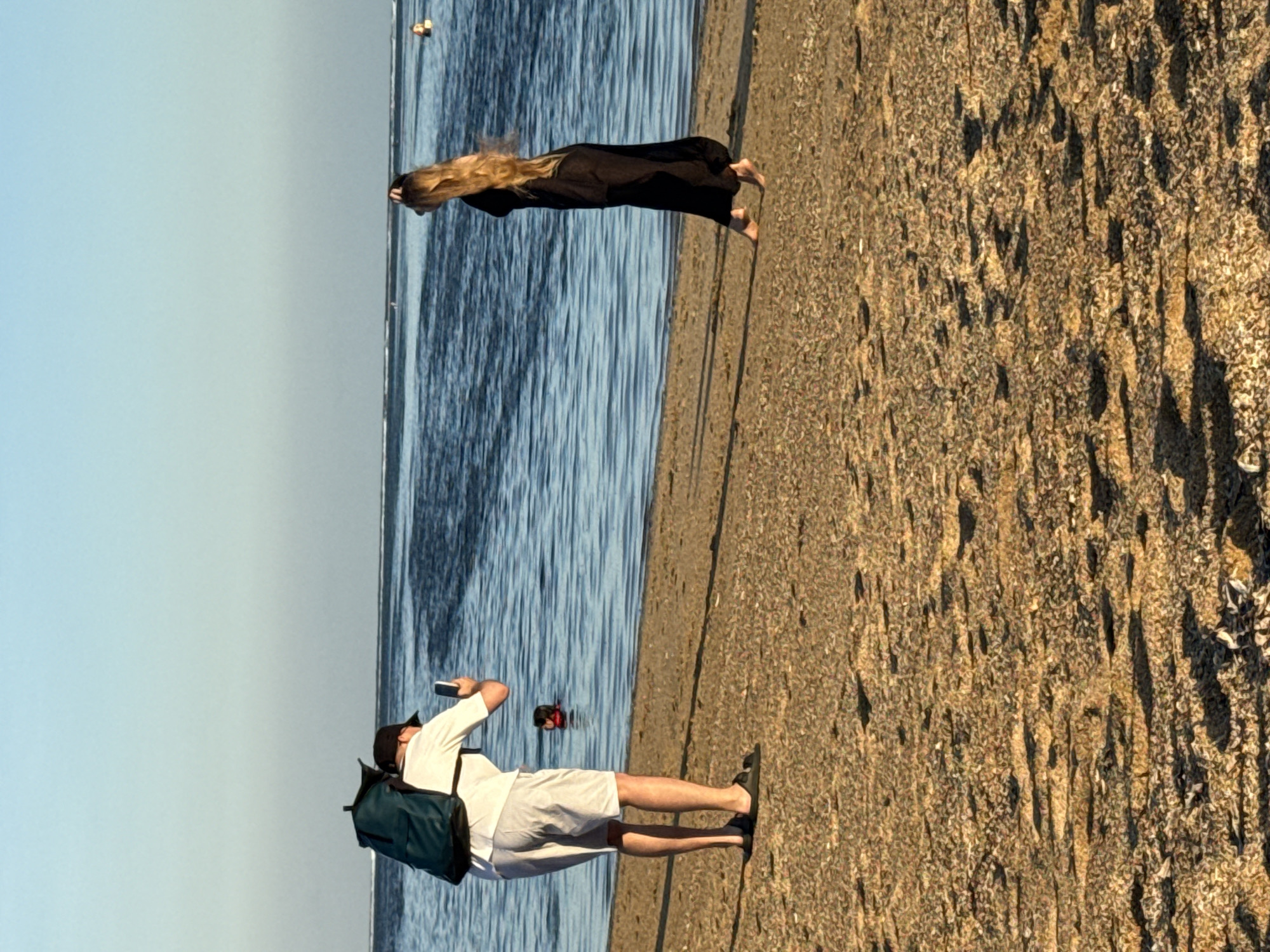 In Denpasar, Indonesia, a man wearing a white t-shirt, grey shorts, a black baseball cap, and a teal backpack is standing on a sandy beach, facing the ocean and holding a smartphone to take a photo. He is positioned in the foreground, with his back to the viewer. In the mid-ground, a woman with long blonde hair, wearing a long black dress, is walking along the shoreline, facing away from the camera. The ocean stretches out behind them, with gentle waves and a clear blue sky above. Several other people are visible in the water in the distance. The beach itself is covered in sand and small pebbles, with some scattered shadows cast by the sunlight.