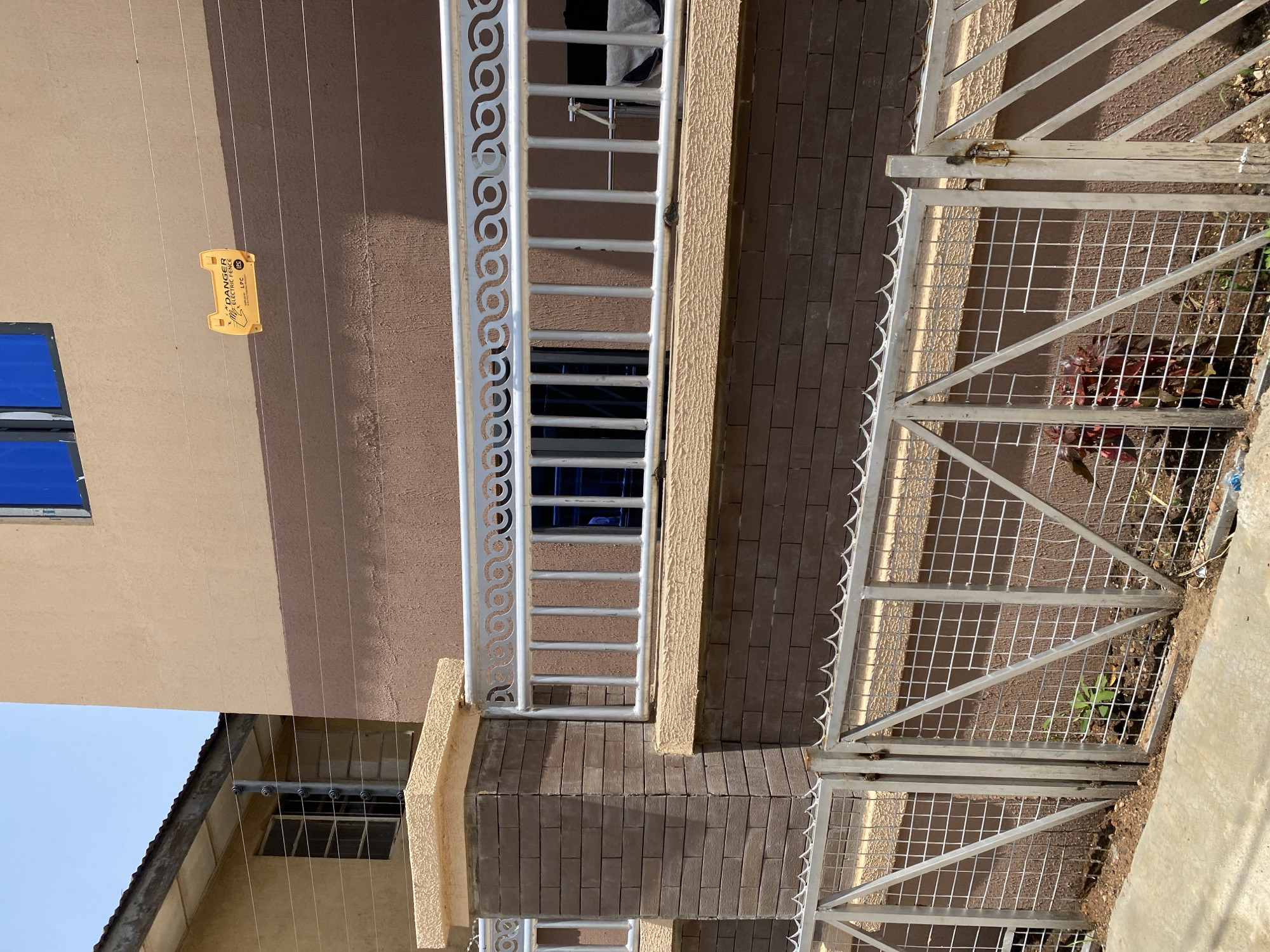 The image depicts the exterior of a building in Jos, Nigeria, showcasing security features. A brown brick wall forms the base of the building, topped with a beige-colored decorative ledge. Above this, a light brown painted wall has a darker brown stripe running horizontally. A prominent feature is an electric fence, consisting of multiple wires, with a yellow "DANGER ELECTRIC FENCE" warning sign attached. Decorative metal railings with an intricate scroll pattern are mounted above a brick pillar and extend along the building's facade. A lower, more utilitarian metal fence with a diamond mesh pattern and pointed spikes along its top separates the property from the paved walkway. The scene is viewed outdoors under a bright, clear sky.