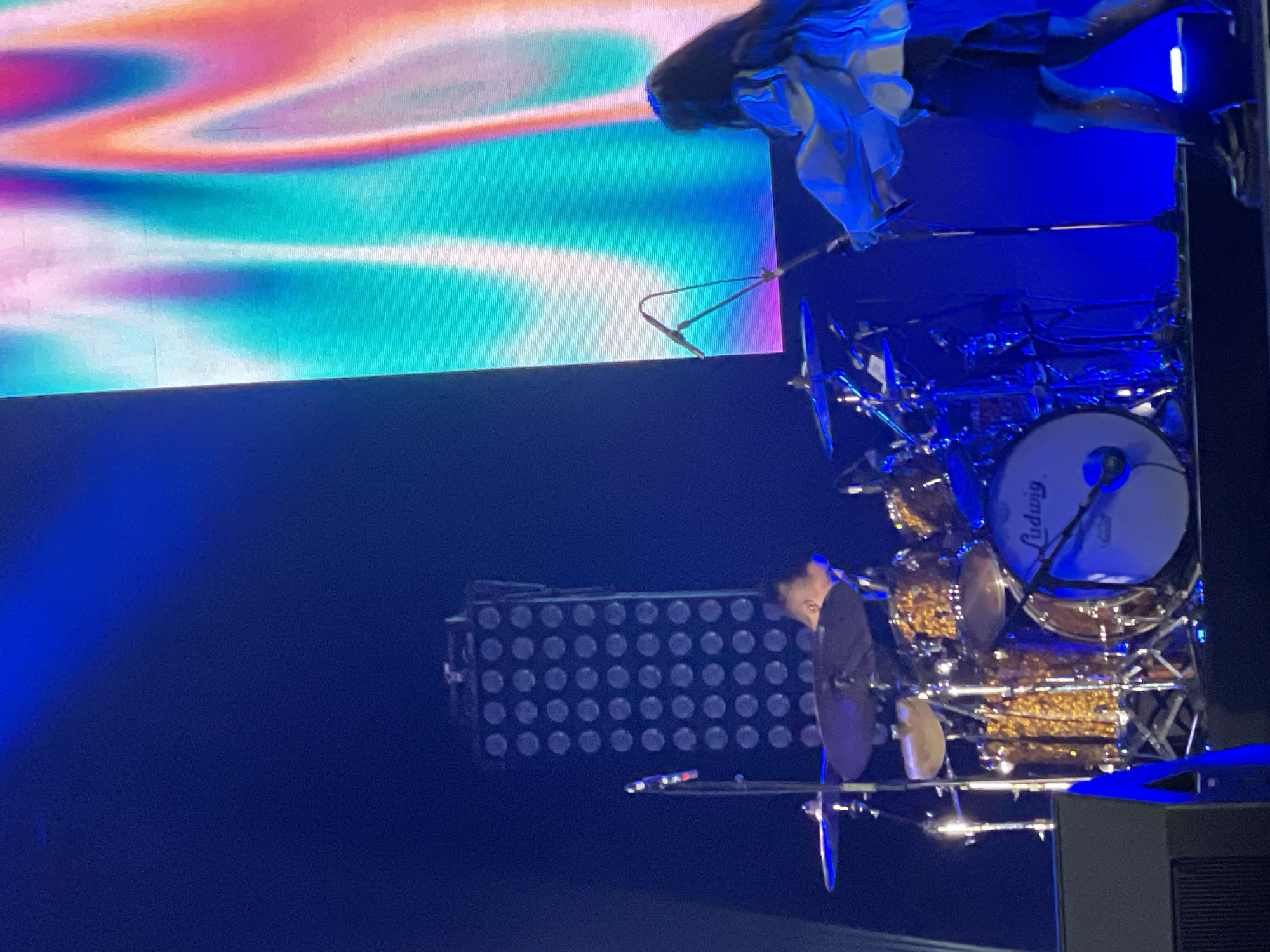 The image shows a drummer performing on a stage in front of a large screen. The screen displays a colorful, abstract pattern. The drummer is positioned behind a drum kit. A lighting rig is also visible on the stage. It is unknown whether the location is Salembaran, Indonesia.
