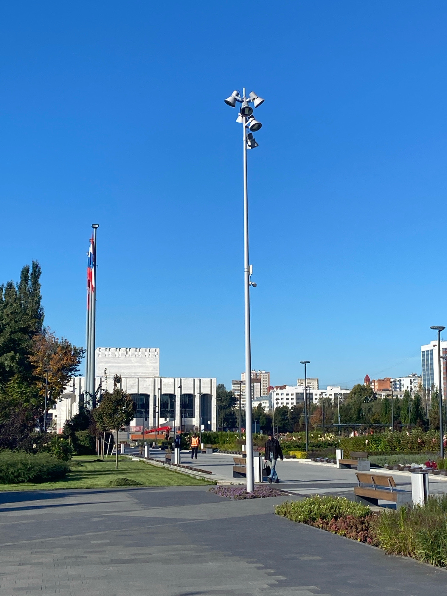 The image depicts a sunny day in a modern urban park in Perm, Russia. In the foreground, a wide paved pathway, constructed with dark gray rectangular pavers, leads towards a landscaped area. To the right, several wooden park benches are visible, some occupied by individuals. A person in a dark jacket and jeans walks along the pathway, carrying a bag. To the left, a lush green lawn is bordered by various plants and trees, some displaying autumn colors.
In the mid-ground, a tall, slender lamppost with multiple spotlights stands prominently, illuminated against the clear blue sky. Further back, a grand building with a white facade and large windows is situated, possibly a cultural or administrative center. Two flags, one predominantly red and white, and another with red, white, and blue stripes, are hoisted on tall flagpoles to the left of the building. The background is filled with the cityscape of Perm, featuring numerous buildings of varying heights, interspersed with trees. The overall scene conveys a sense of urban development and public amenity in Perm.