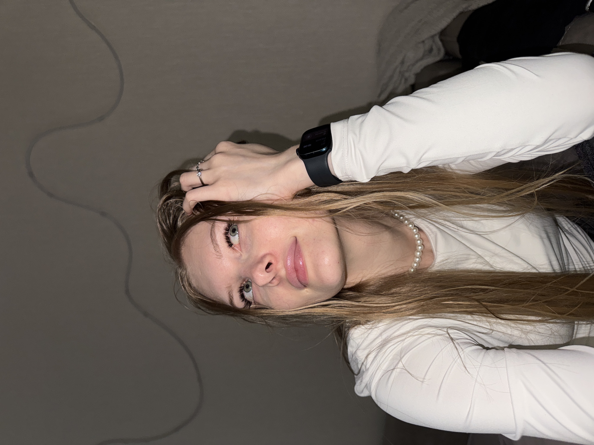 The image displays a young woman in an indoor setting. She has long, light brown hair, light-colored eyes, and is wearing a white long-sleeved top and a pearl necklace. A silver ring adorns her left ring finger, and a black smartwatch is visible on her left wrist. The smartwatch screen clearly shows the time as "16:06" and includes text such as "Health".
Her left hand is positioned near her head, and she looks upwards with a gentle, slightly pensive expression, her lips glossy pink. The background is a plain, dark gray wall with a subtle, wavy line pattern. The lighting in the scene appears dim, indicating an indoor environment possibly at dusk or in a low-light room. The photograph is oriented sideways, with the subject's head on the right side of the frame. This scene takes place in Perm, Russia.