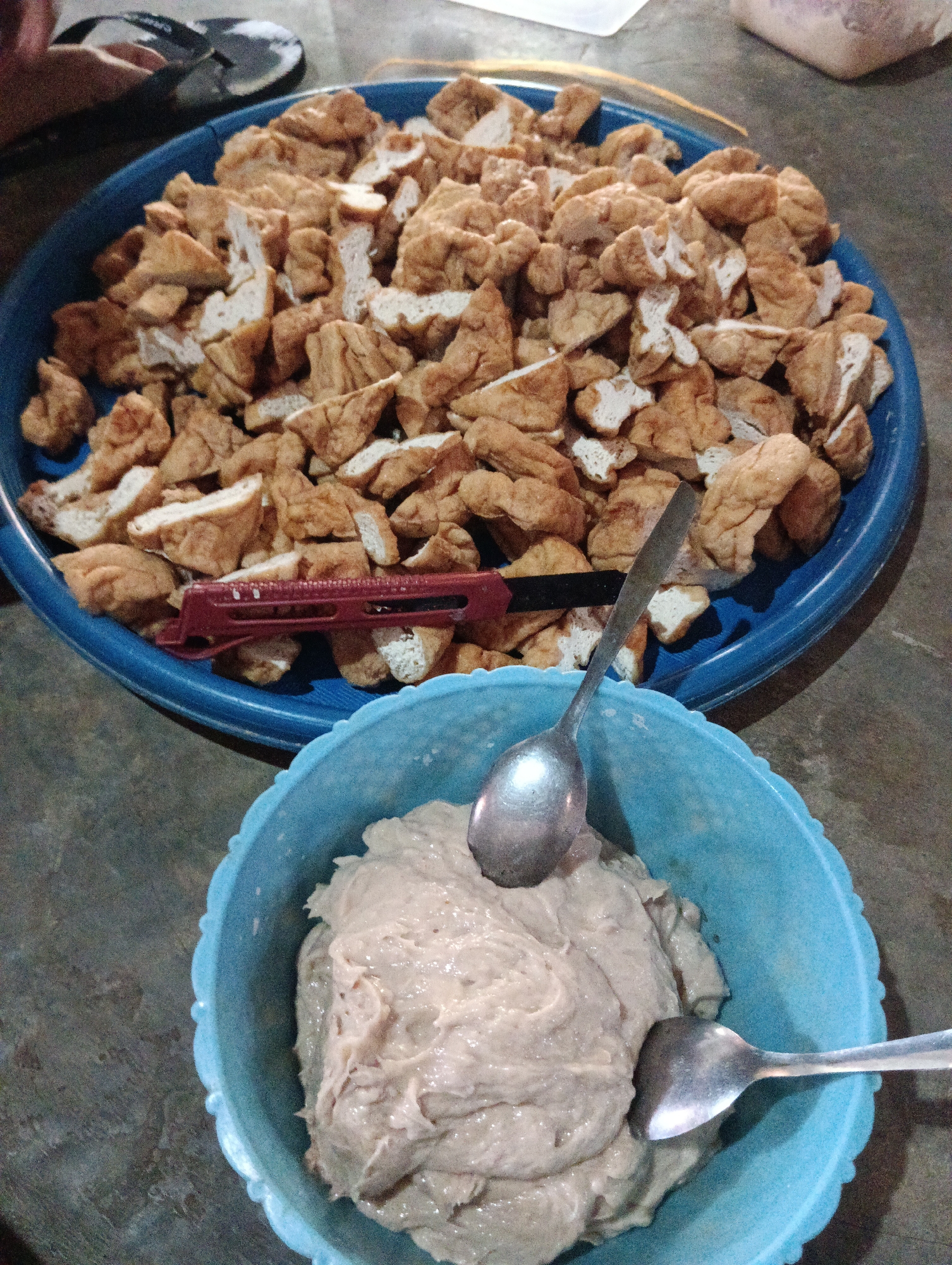 In Pematang Jaya, Indonesia, a close-up shot shows a blue plastic bowl filled with a pale, creamy mixture, likely a food preparation. Two metal spoons are placed in the mixture. In the background, a larger blue plastic basin contains numerous pieces of fried tofu, cut into bite-sized chunks. A red and black handled utensil, possibly a small knife or tongs, rests on top of the tofu. The scene appears to be indoors, with a textured, concrete-like floor. A portion of a person's hand and a black sandal are visible in the upper left corner.