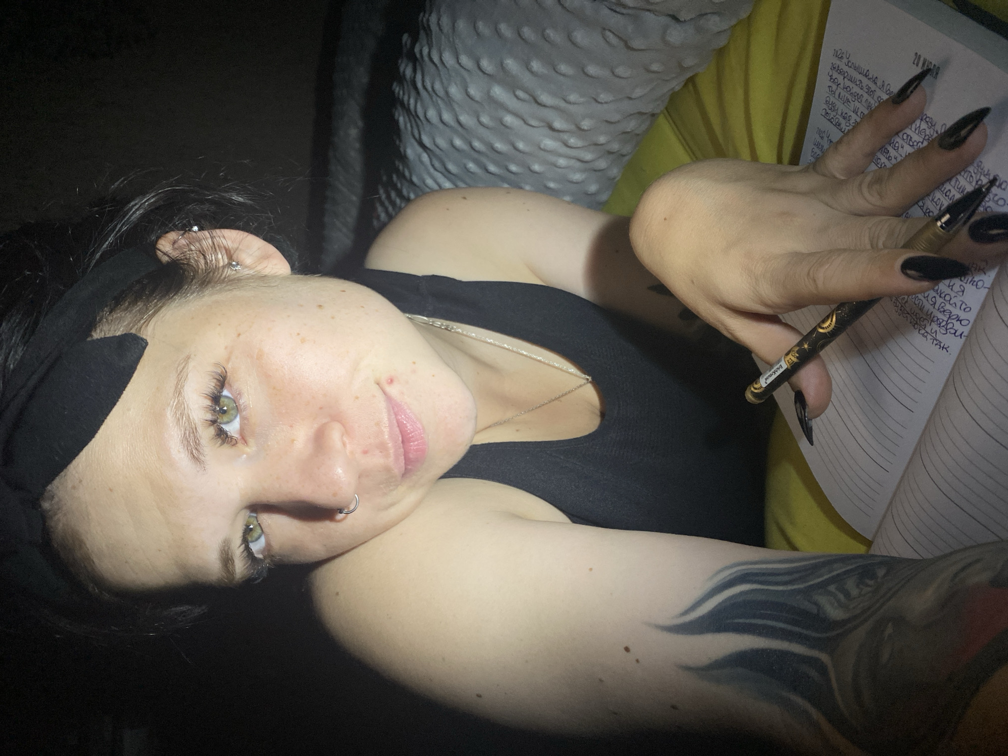 The image shows a woman lying on a bed in a dimly lit room. She is wearing a black tank top and a black headband. She has long, dark hair and green eyes, and she is wearing a nose ring and earrings. She has a tattoo on her left arm and long, pointed black nails. She is holding a pen and writing in a notebook. The room appears to be a bedroom, with a pillow and a yellow sheet visible in the background. The lighting suggests it is nighttime. There is no information to suggest this is in Chaikovsky, Russia.
