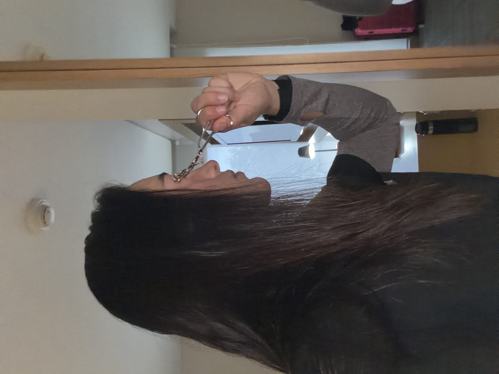 A woman with long, dark hair is using an eyelash curler on her right eye. She is wearing a dark shirt with a grey sleeve. The scene appears to be an indoor setting, possibly a room with a mirror, as a reflection of the woman and the room is visible. A pink suitcase and a black thermos are also present in the background. There is no information in the image to suggest it is located in Bogota, Colombia.